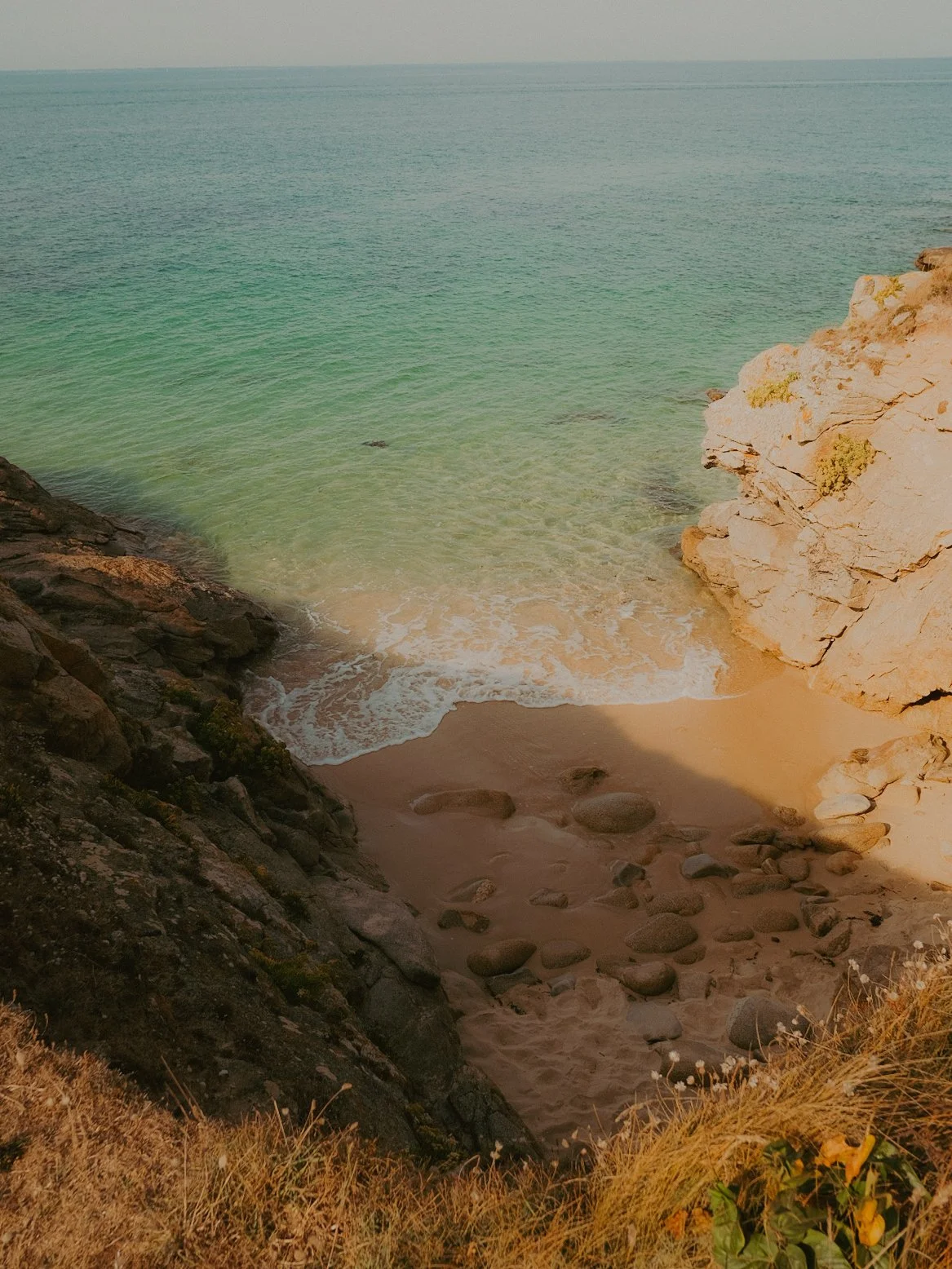 Vue d'une petite plage rocheuse avec des eaux calmes et transparentes, entourée de falaises rocheuses et de végétation sèche en premier plan.