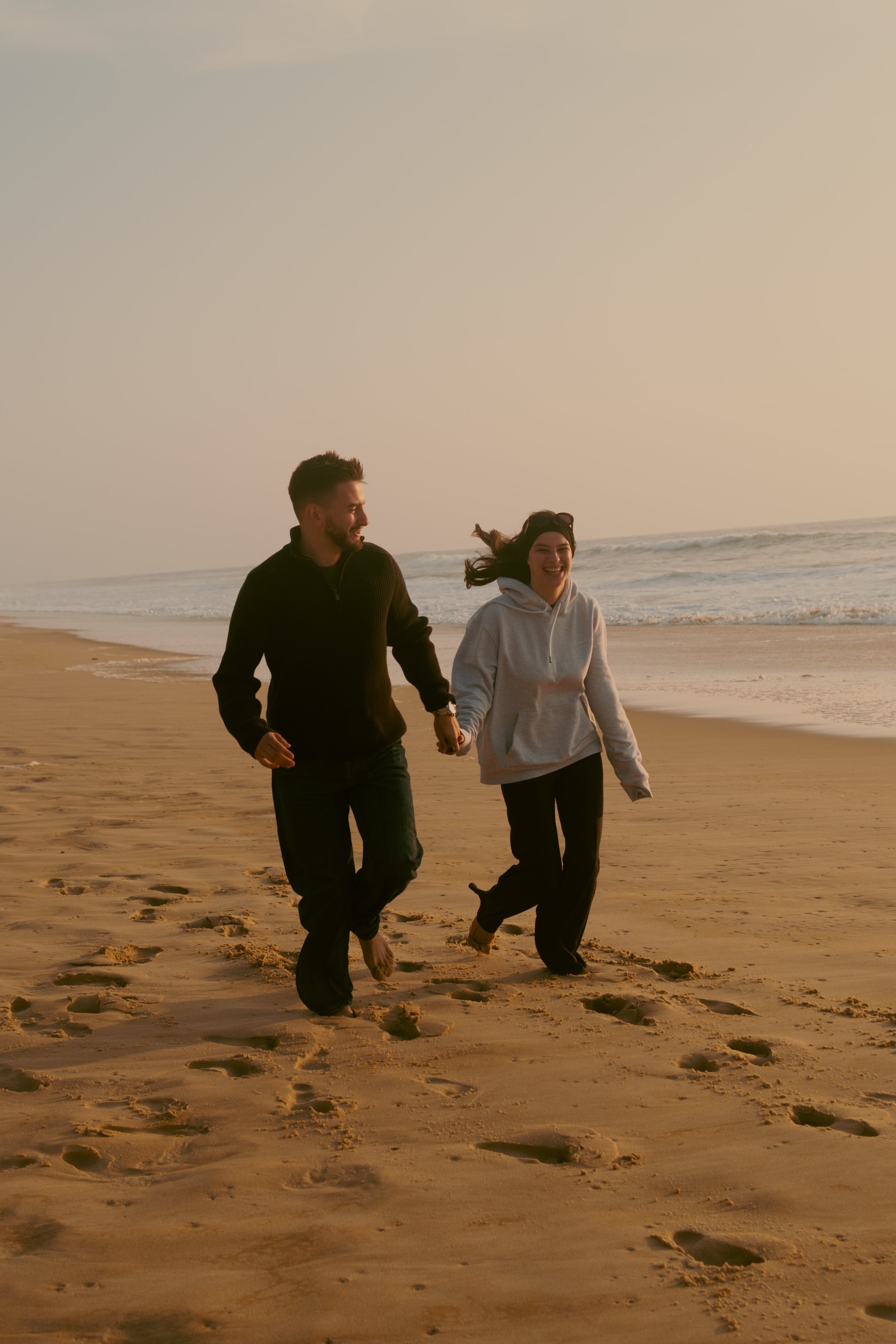 Un couple marche main dans la main sur la plage au coucher du soleil, souriants et heureux.