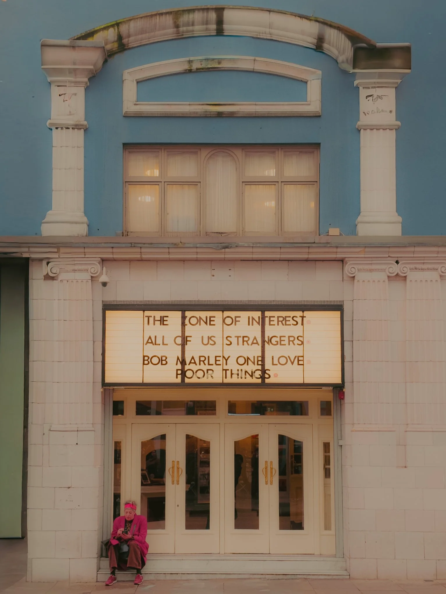 Façade d'un bâtiment blanc avec un panneau lumineux affichant une citation, une femme en vêtements roses assise en bas devant une porte en verre.