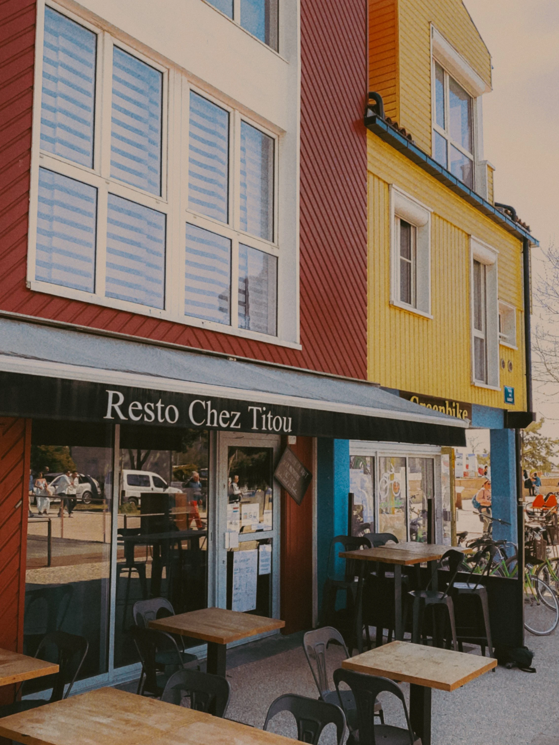Façade d'un restaurant appelé 'Resto Chez Titou' avec des tables et des chaises à l'extérieur, bâtiment coloré en rouge, jaune, et bleu, et fenêtres avec volets.