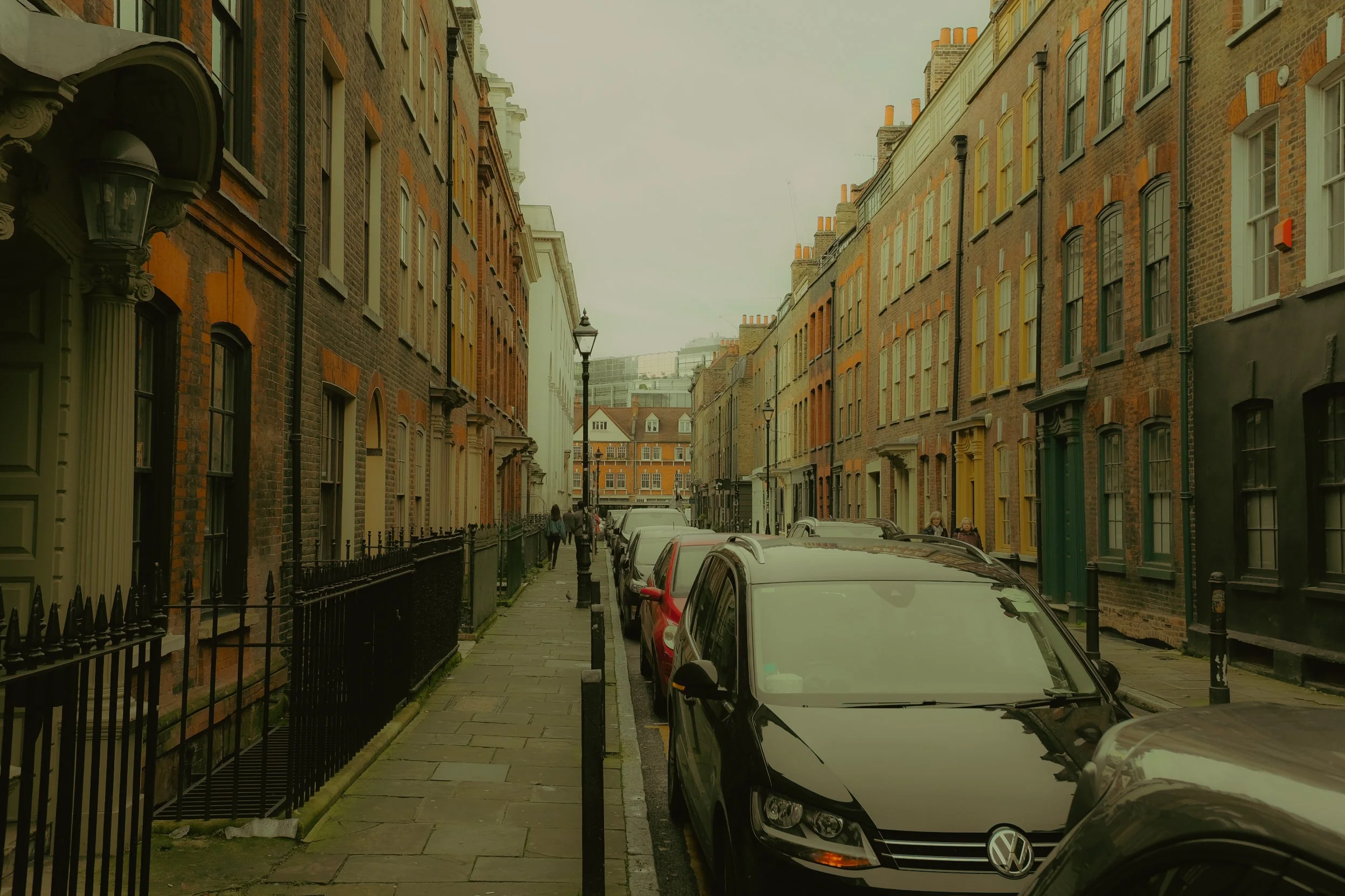 Rue urbaine pavée bordée de bâtiments en briques, avec des voitures garées sur le côté et des lampadaires. Un groupe de personnes marche le long du trottoir.