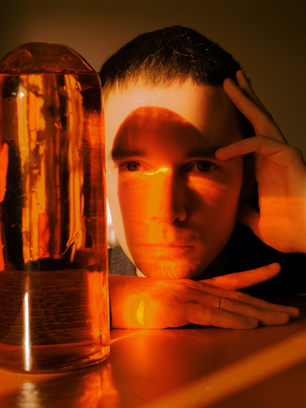 Un jeune homme repose sa tête et ses bras sur une table en regardant fixement vers l'objectif avec une expression contemplative, une lampe à lumière douce éclaire la scène et une grande cuve en verre contenant une boisson orange est visible à côté de