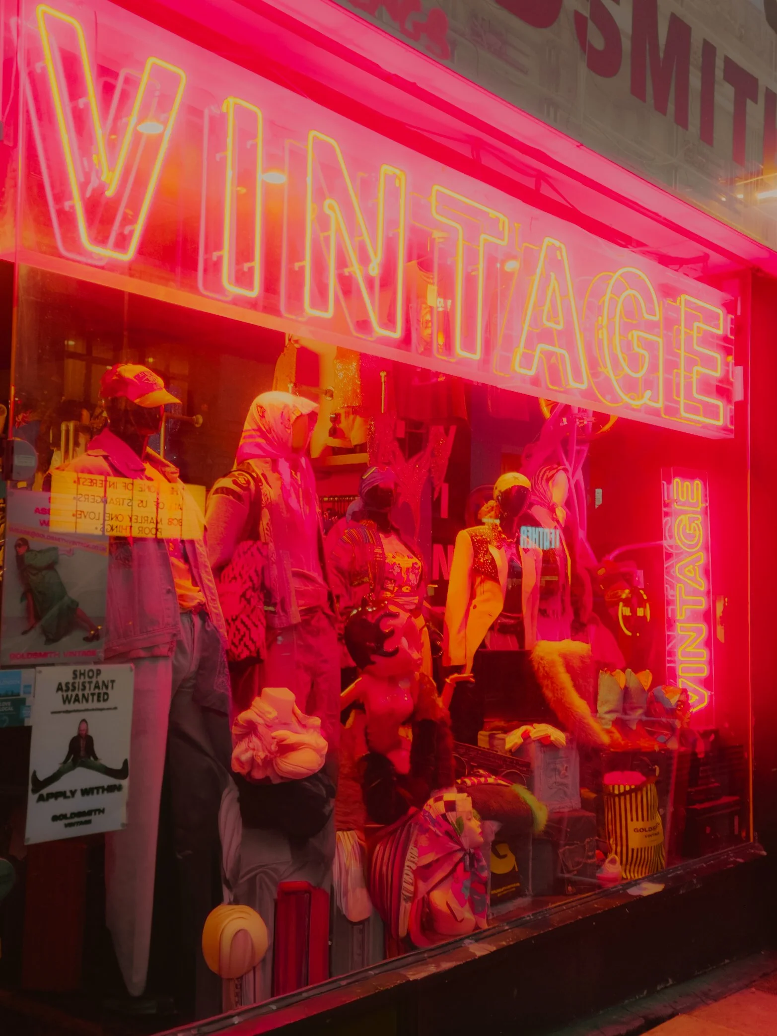 Vitrine d'une boutique avec un affichage lumineux en lettres roses indiquant 'VINTAGE', avec des mannequins vêtus de vêtements vintage et des accessoires en vitrine, le tout illuminé d'une lumière rose.
