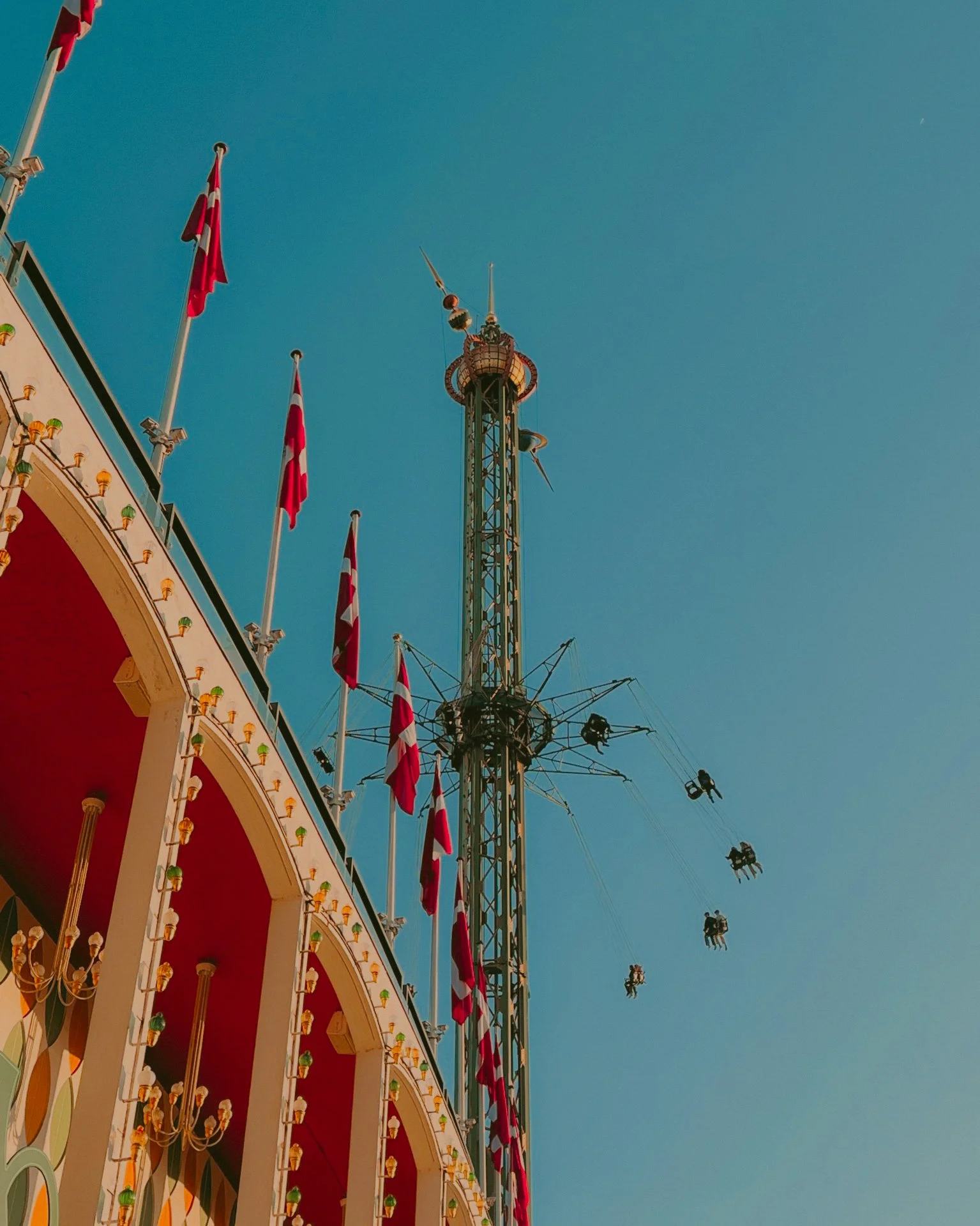 Manège avec plusieurs drapeaux rouges et blancs, structure en acier sous un ciel bleu, avec des sièges suspendus en train de tourner.