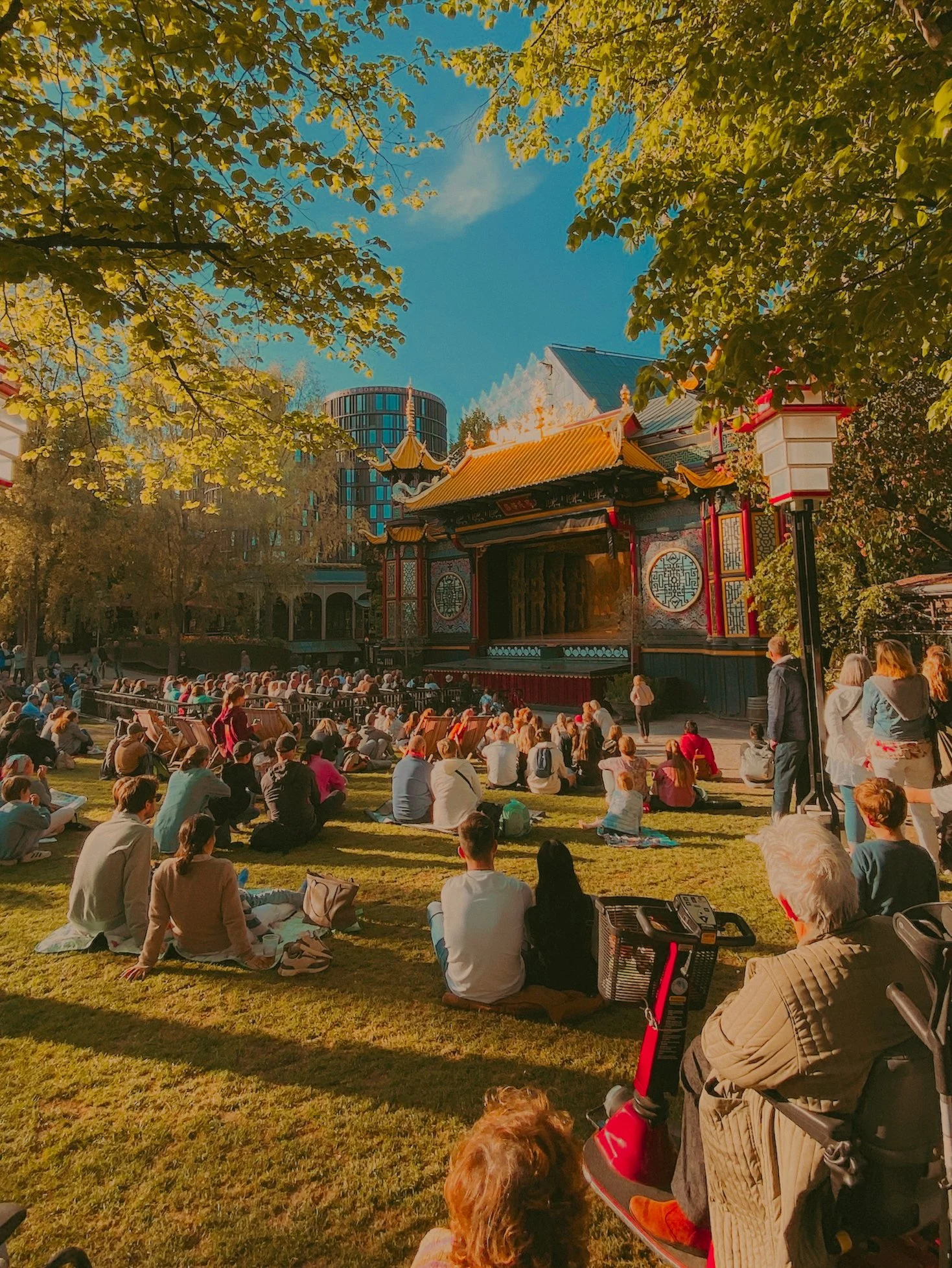 Un groupe de personnes assises sur l'herbe regardant une performance sur scène dans un parc avec des arbres et un bâtiment en fond, sous un ciel ensoleillé.