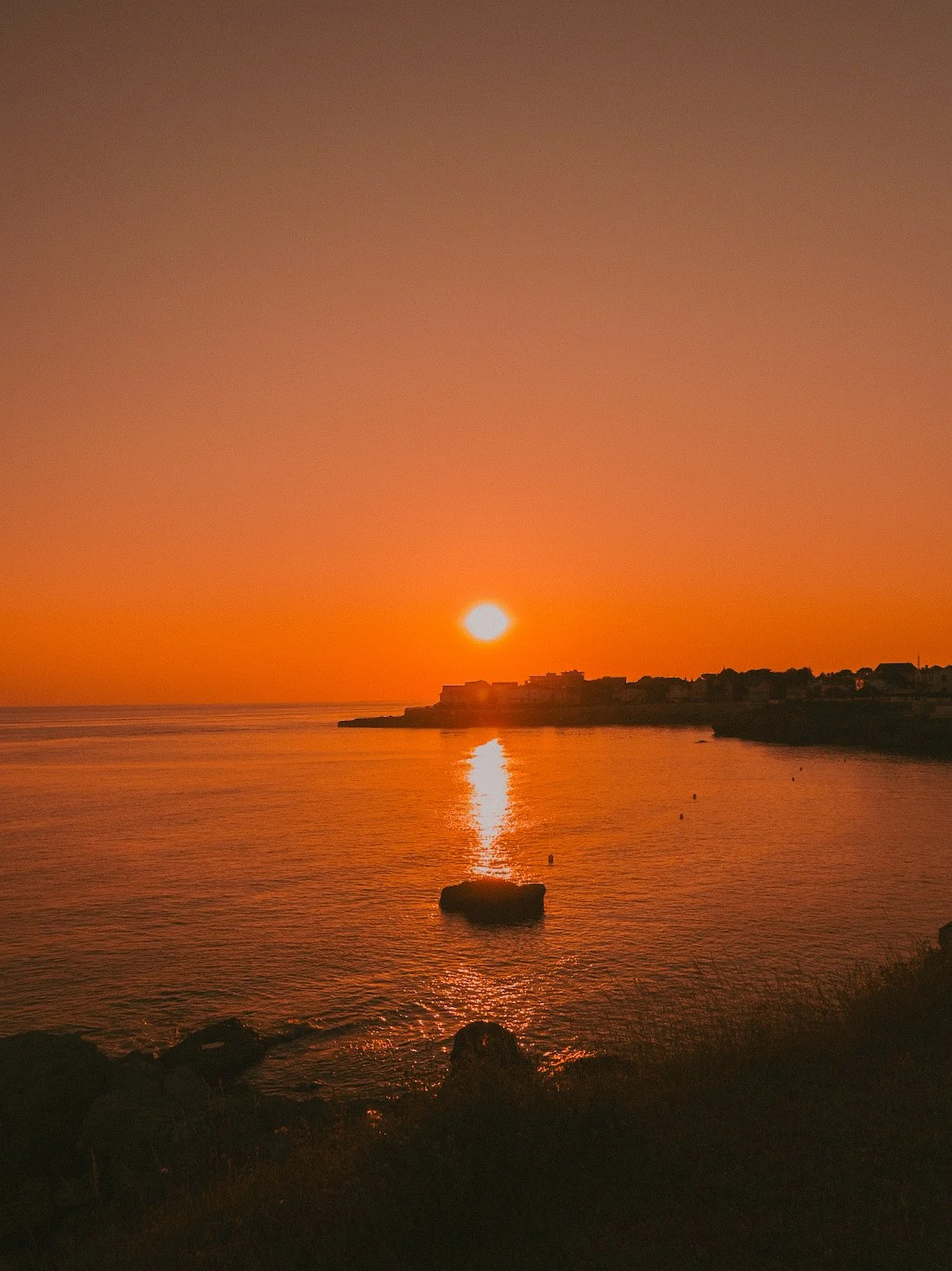 Coucher de soleil sur la mer avec un reflet sur l'eau, horizon avec quelques bâtiments silhouette et un rocher dans l'eau.