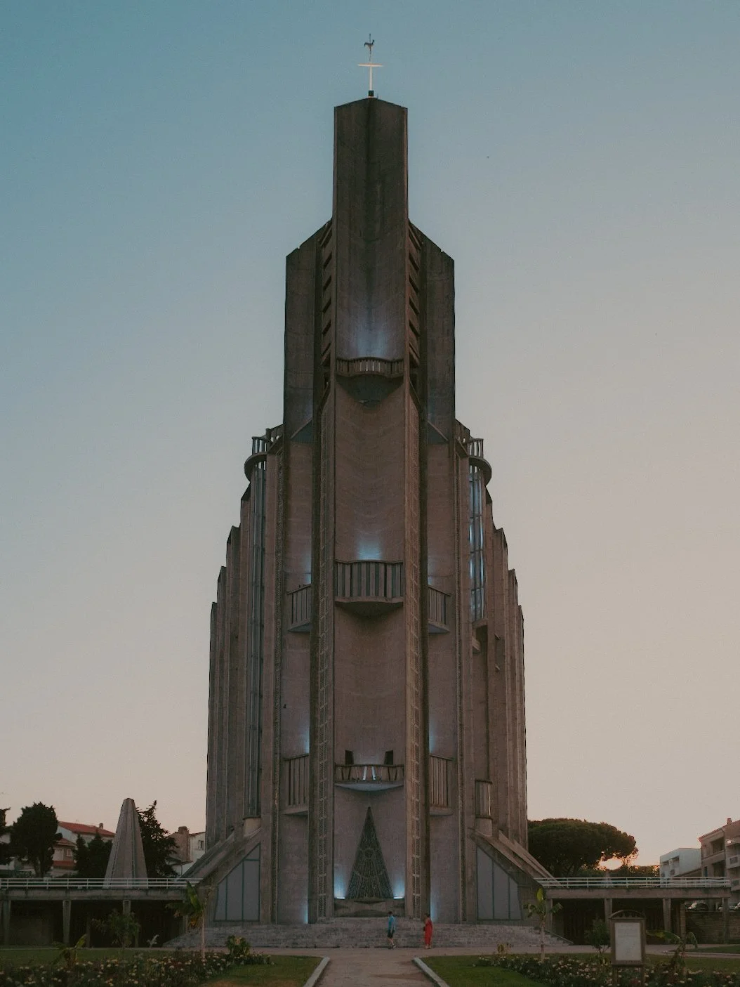 Une grande église ou cathédrale en béton avec un style architectural moderne, vue de face, à l'heure du coucher ou du lever du soleil avec un ciel clair, entourée de petits arbres et de bâtiments résidentiels.