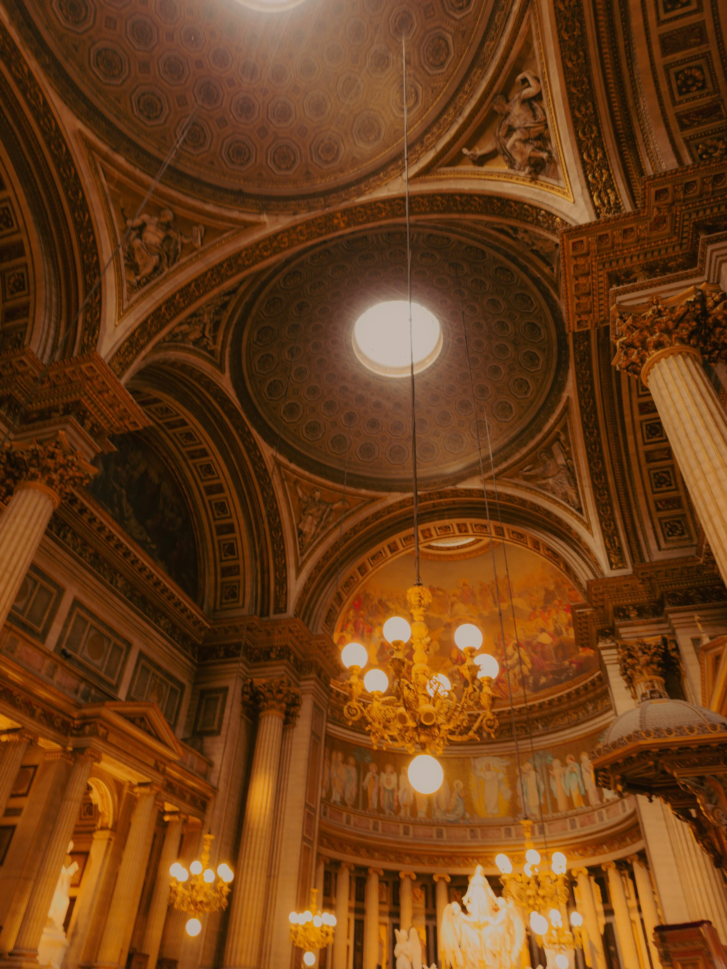 Intérieur d'une cathédrale ou d'une église avec un plafond orné, des colonnes et un lustre chandeliers en or, un oculus au plafond laissant passer la lumière, et des fresques ou peintures religieuses en arrière-plan.