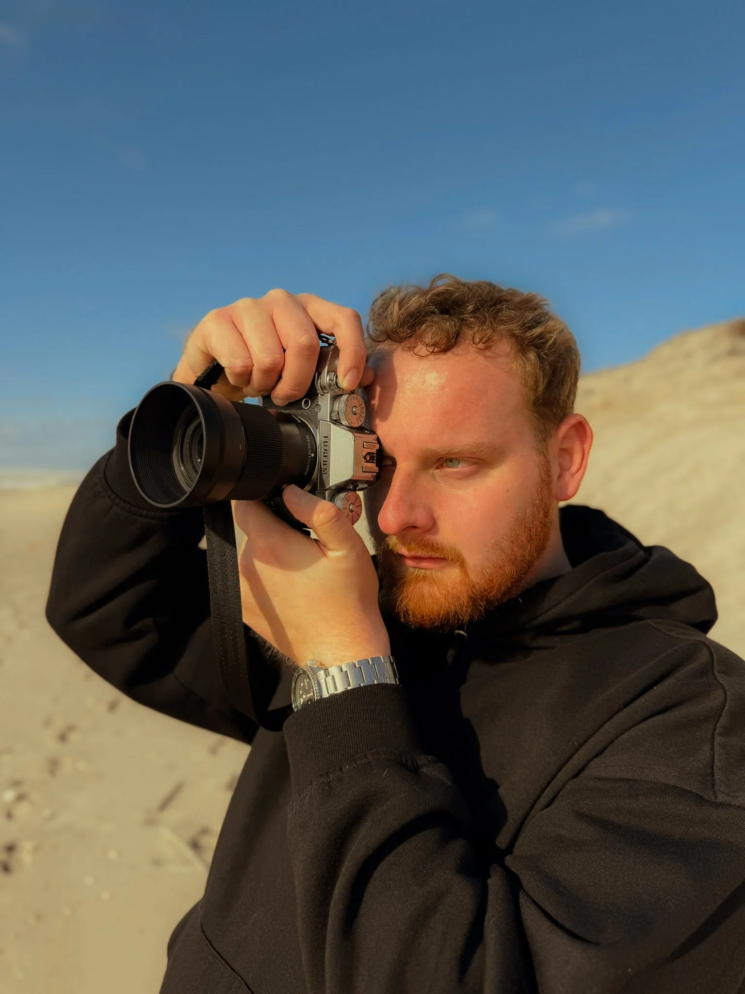 Un homme à la barbe rousse, portant une montre argentée, prend une photo avec un appareil photo numérique lors d'une journée ensoleillée à la plage sur fond de ciel bleu.