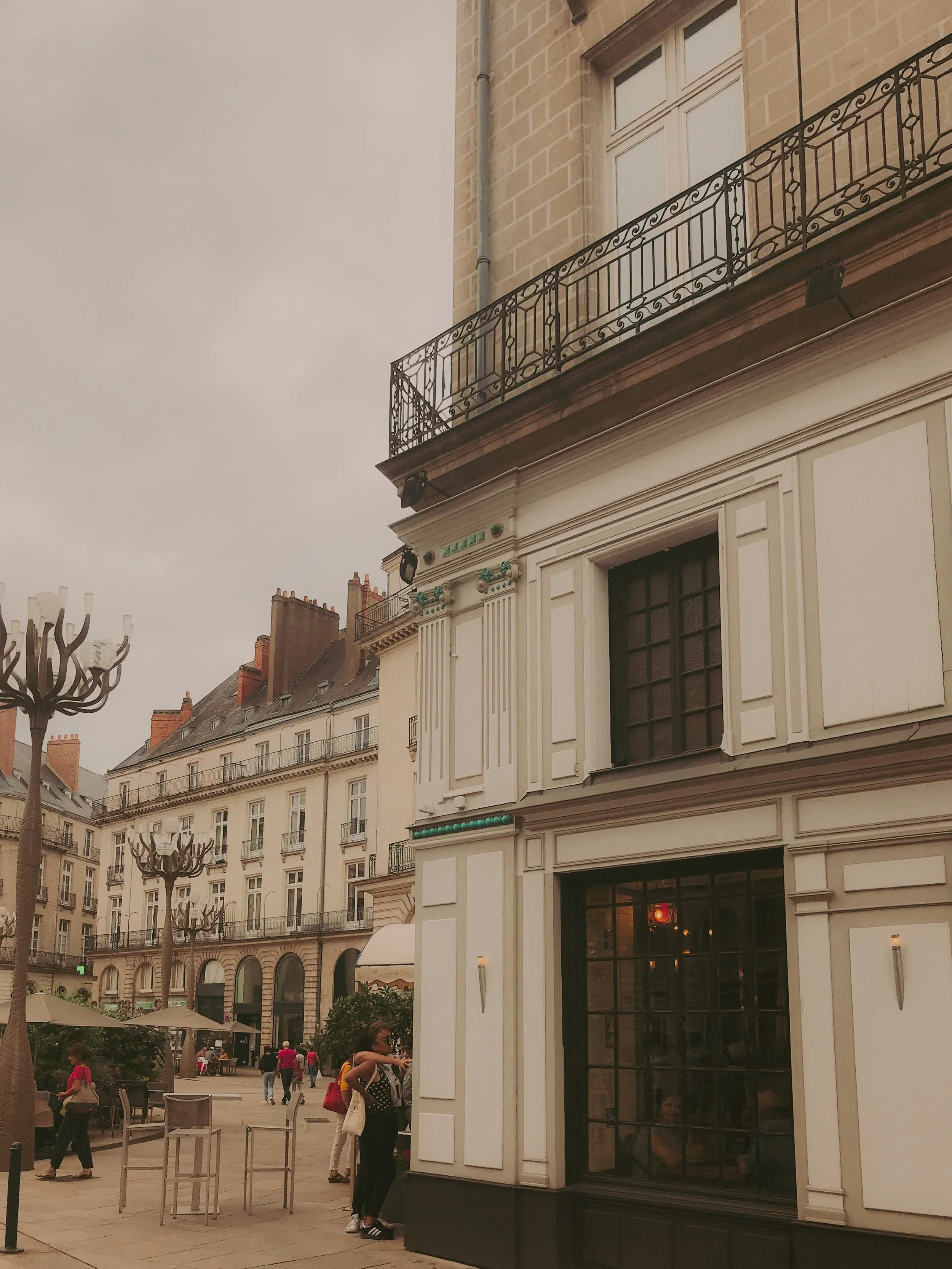 Une vue de bâtiments haussmanniens dans une rue animée parisienne. En premier plan, une femme avec un sac à dos et une femme assise à une fenêtre d'un café. Des lampadaires en forme d'arbres sont aussi visibles.