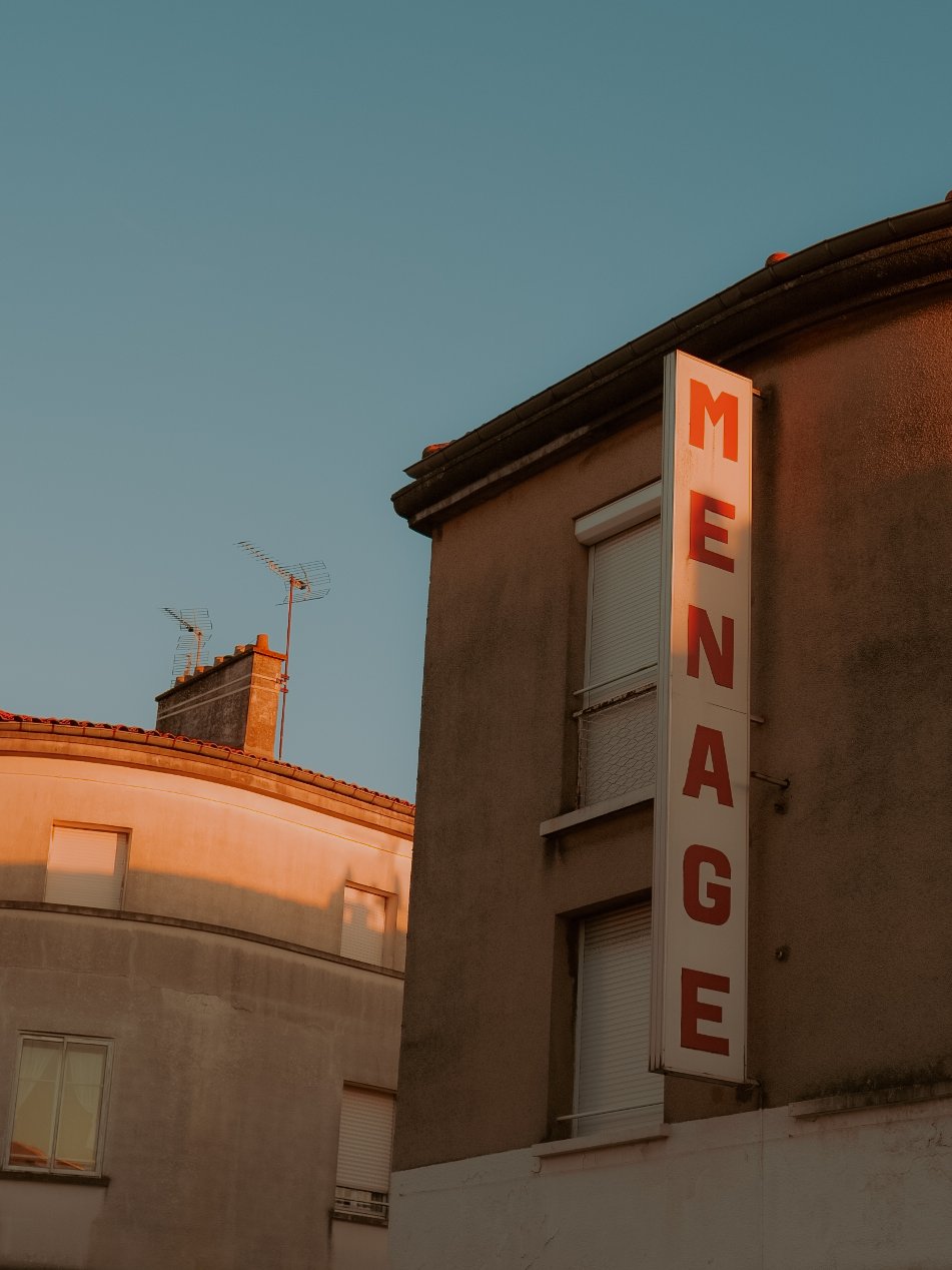 Un panneau vertical affiche le mot « MENAGE » en grandes lettres rouges sur fond blanc, fixé à un bâtiment gris, sous un ciel clair, avec une autre bâtisse en arrière-plan.