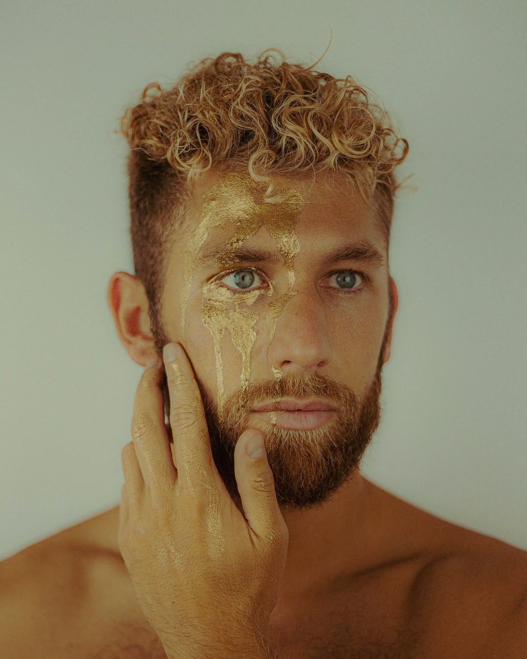 Un homme avec une barbe et des cheveux bouclés, le visage peint en or, touche son visage avec sa main également recouverte de peinture dorée.