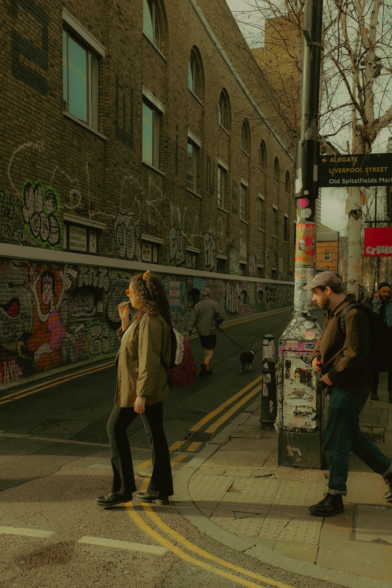 Une scène de rue urbaine avec plusieurs piétons, un chien, un mur recouvert de graffitis et des panneaux de signalisation indiquant Aldgate, Liverpool Street, et Old Spitalfields Market.