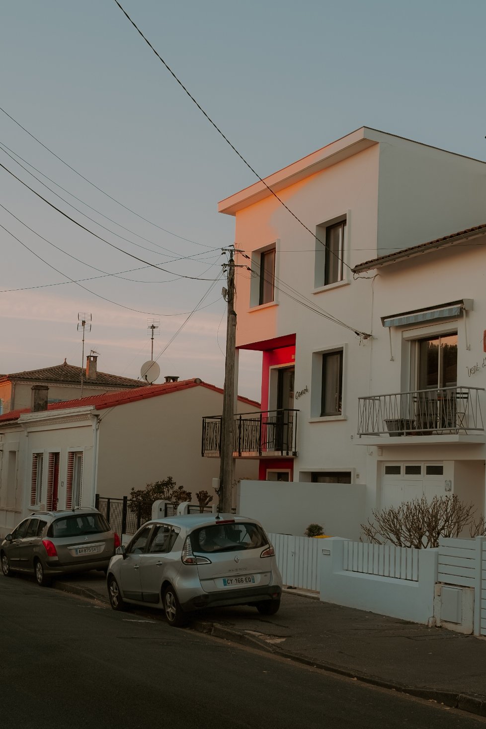 Vue d'une rue résidentielle avec des maisons modernes, des voitures stationnées, un poteau électrique et un ciel au coucher du soleil.