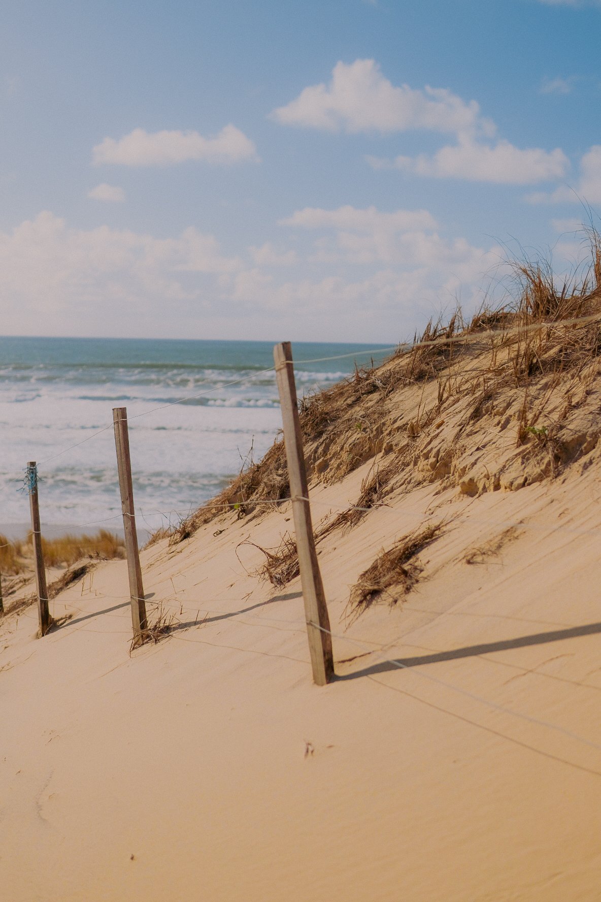 Plage de sable avec des dunes, du grillage et un ciel bleu avec quelques nuages.