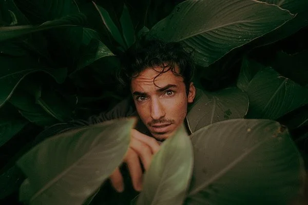 Un homme avec des cheveux bouclés foncés parmi de grandes feuilles vertes, regardant vers la caméra.