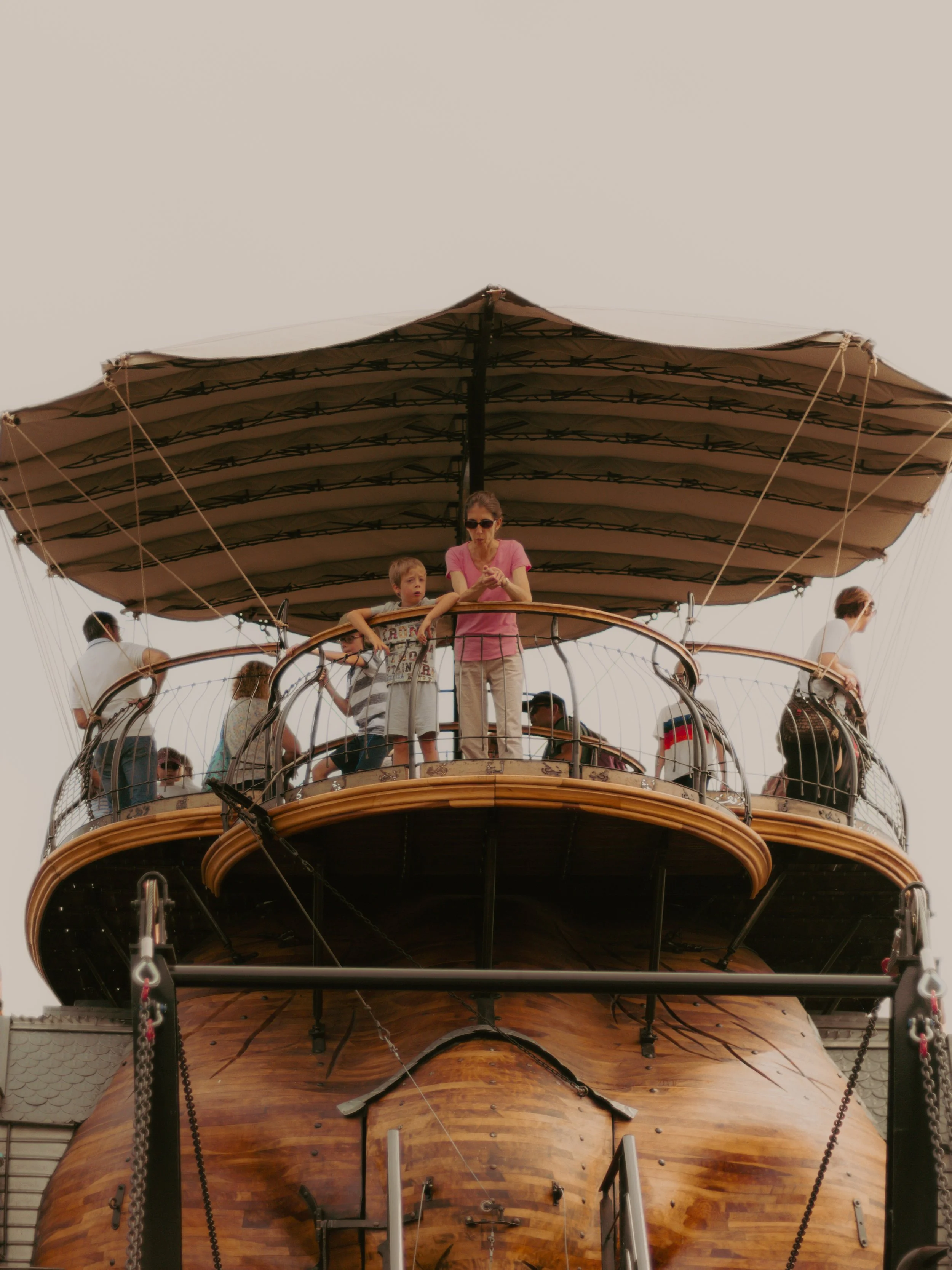 Un bateau en bois avec un grand mât, où plusieurs personnes, dont un adulte et deux enfants, se tiennent sur le pont, avec un ciel clair en arrière-plan.