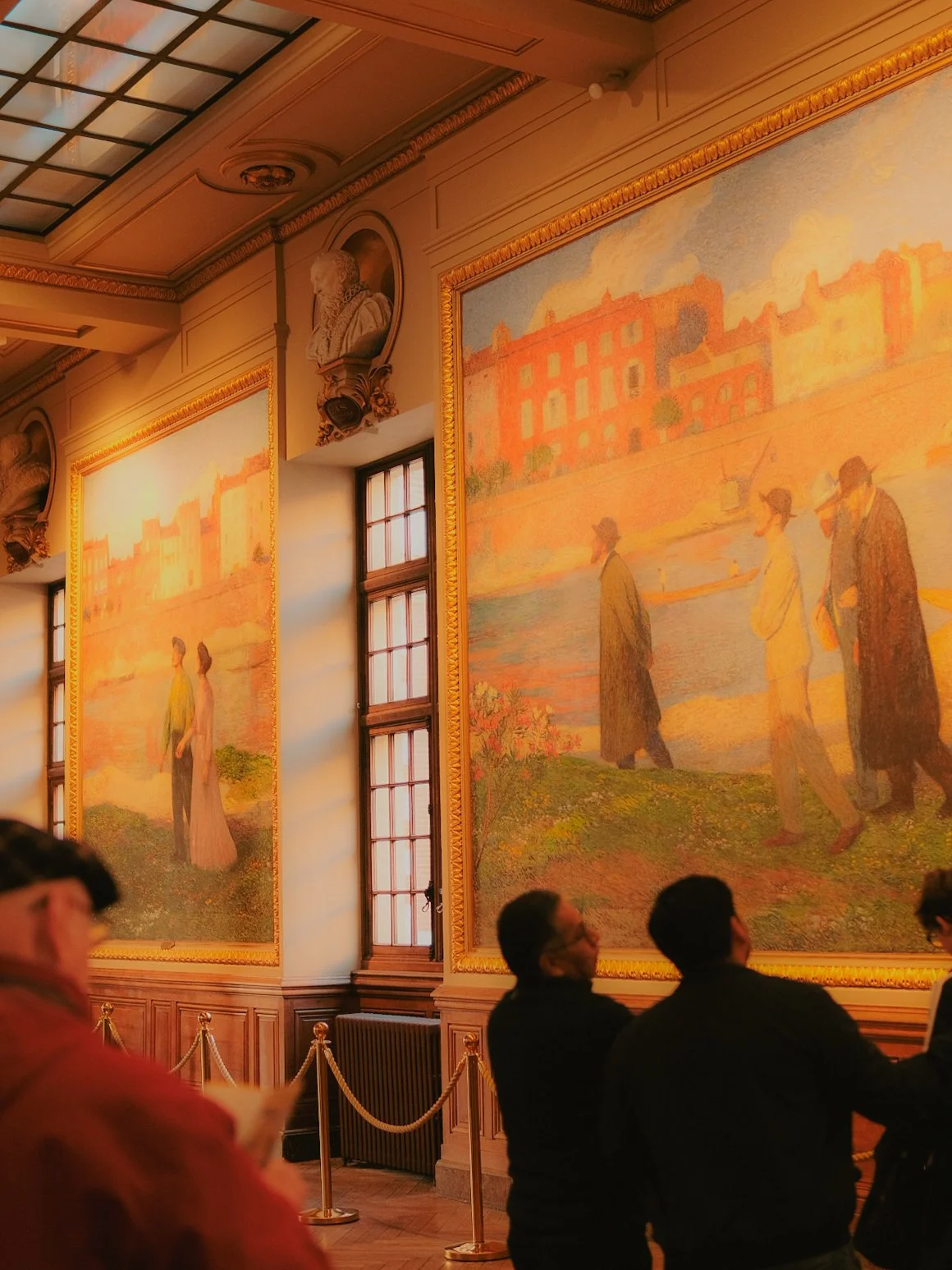 Une salle d'exposition avec de grandes peintures impressionnistes représentant des scènes de navires et de paysages au bord de l'eau. Des visiteurs sont en train d'admirer les œuvres, entourées de décorations anciennes et de moulures dorées.