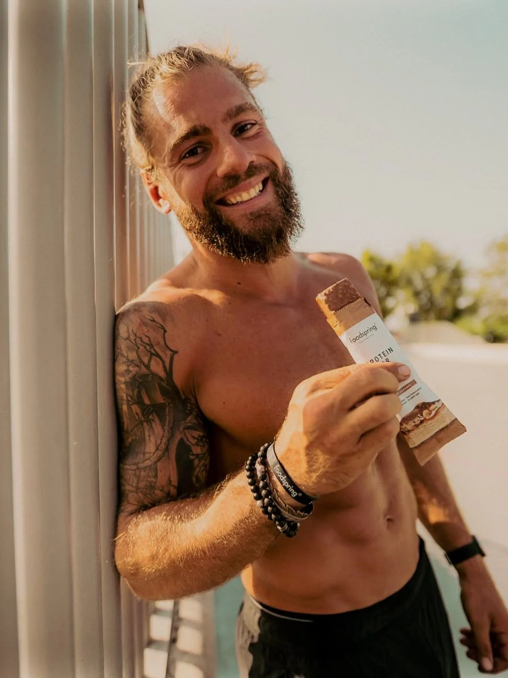 Un homme torse nu avec tatouage sur le bras, souriant, tient une barre énergétique à la main, avec en arrière-plan un ciel clair et des arbres.
