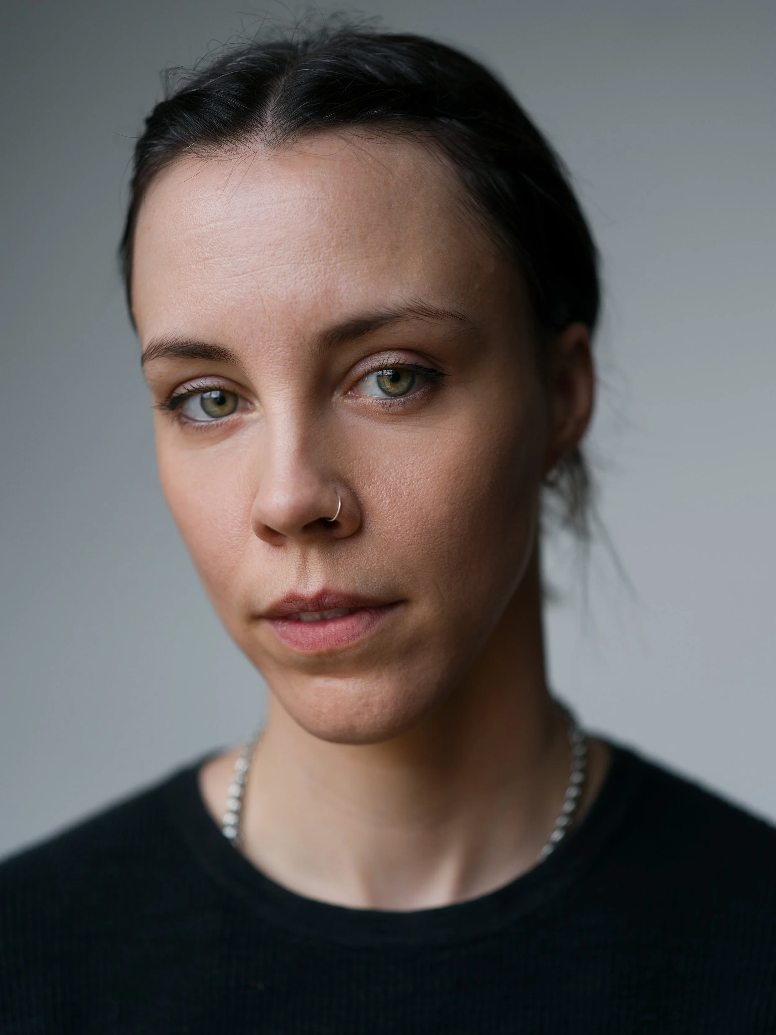 Close-up portrait of a woman with dark hair, green eyes, a nose ring, wearing a black top and silver chain necklace.