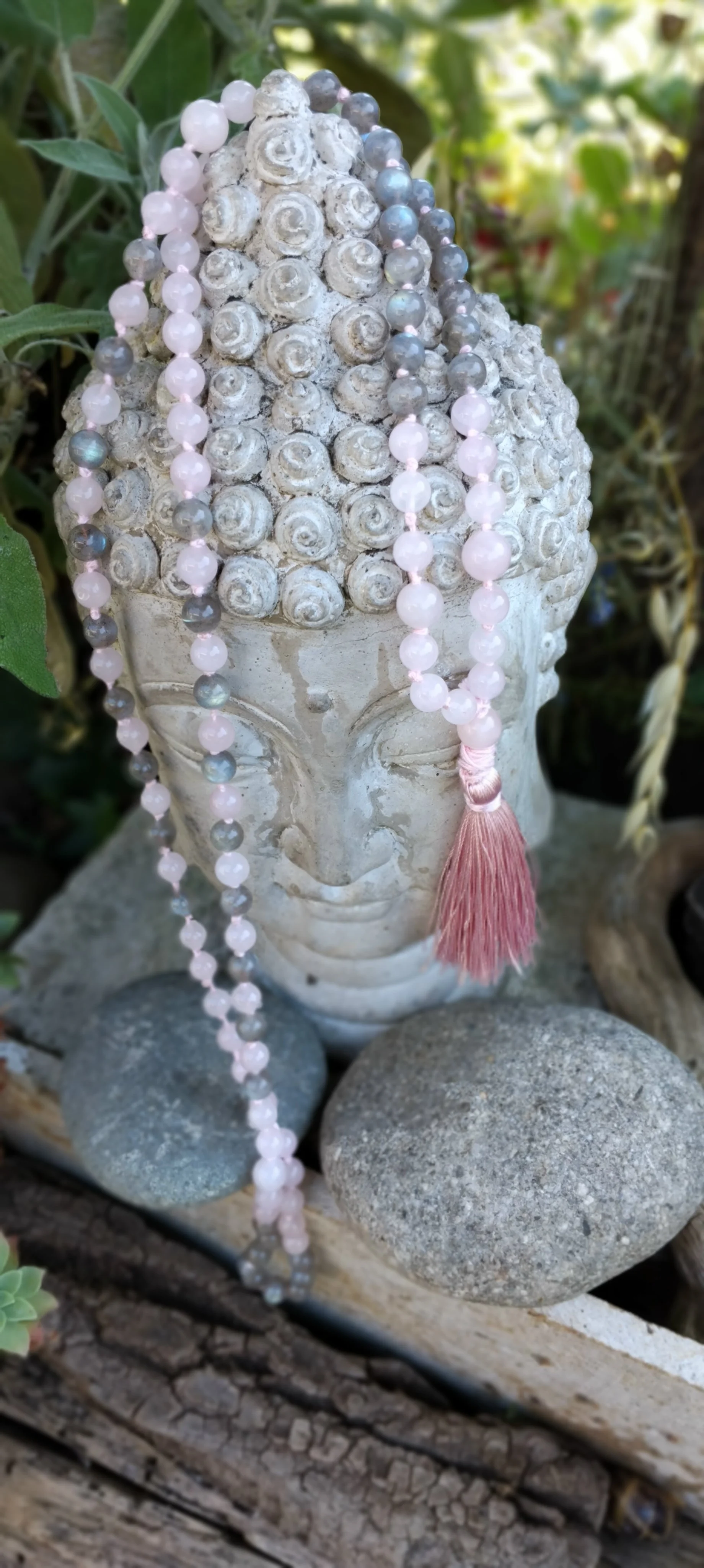 Une tête de sculpture en pierre de style Bouddha ornementée de beads roses et gris, avec un pompon rose suspendu.
