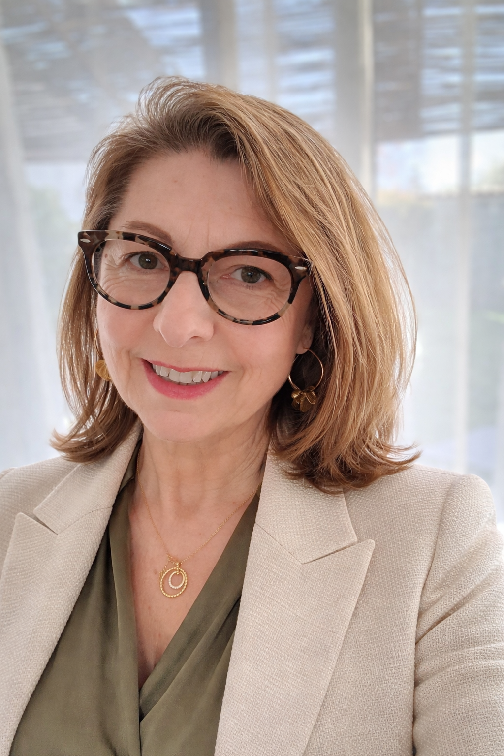 Une femme souriante portant des lunettes à monture tortoise, des boucles d'oreilles dorées, un collier en or et une veste beige, posant devant une fenêtre avec des rideaux lumineux.
