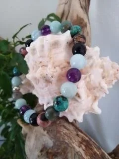 Bracelet chemin de vie en pierres semi précieuses colorées posé sur un corail blanc avec des branches de bois et des feuilles vertes en arrière-plan.
