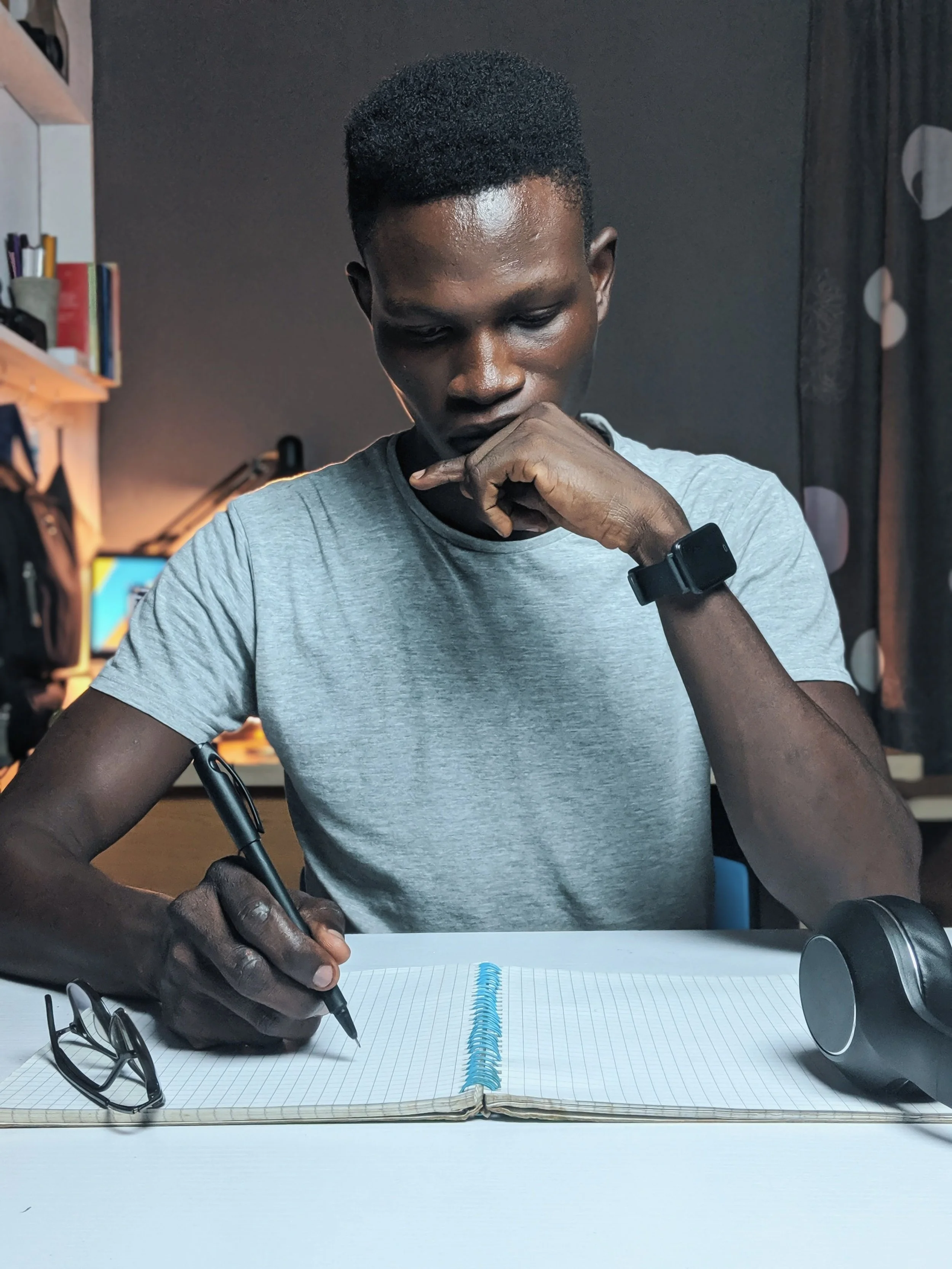 man sitting, wearing white tshirt, with notebook and pen thinking about what to write with headphone and glasses on table