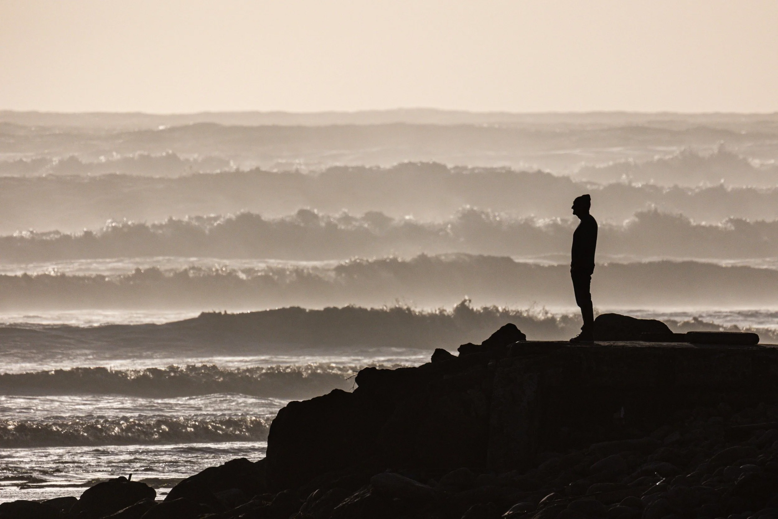 person silhouette wearing a beanie standing on a cliff by the ocean and all its' waves crashing
