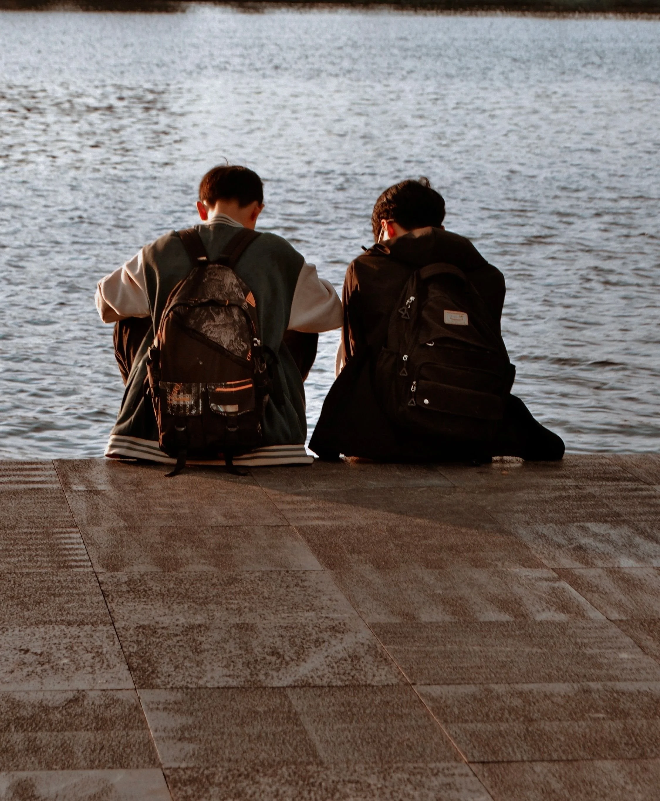two males with jackets and backpacks sitting side by side at a river bank with back facing to camera