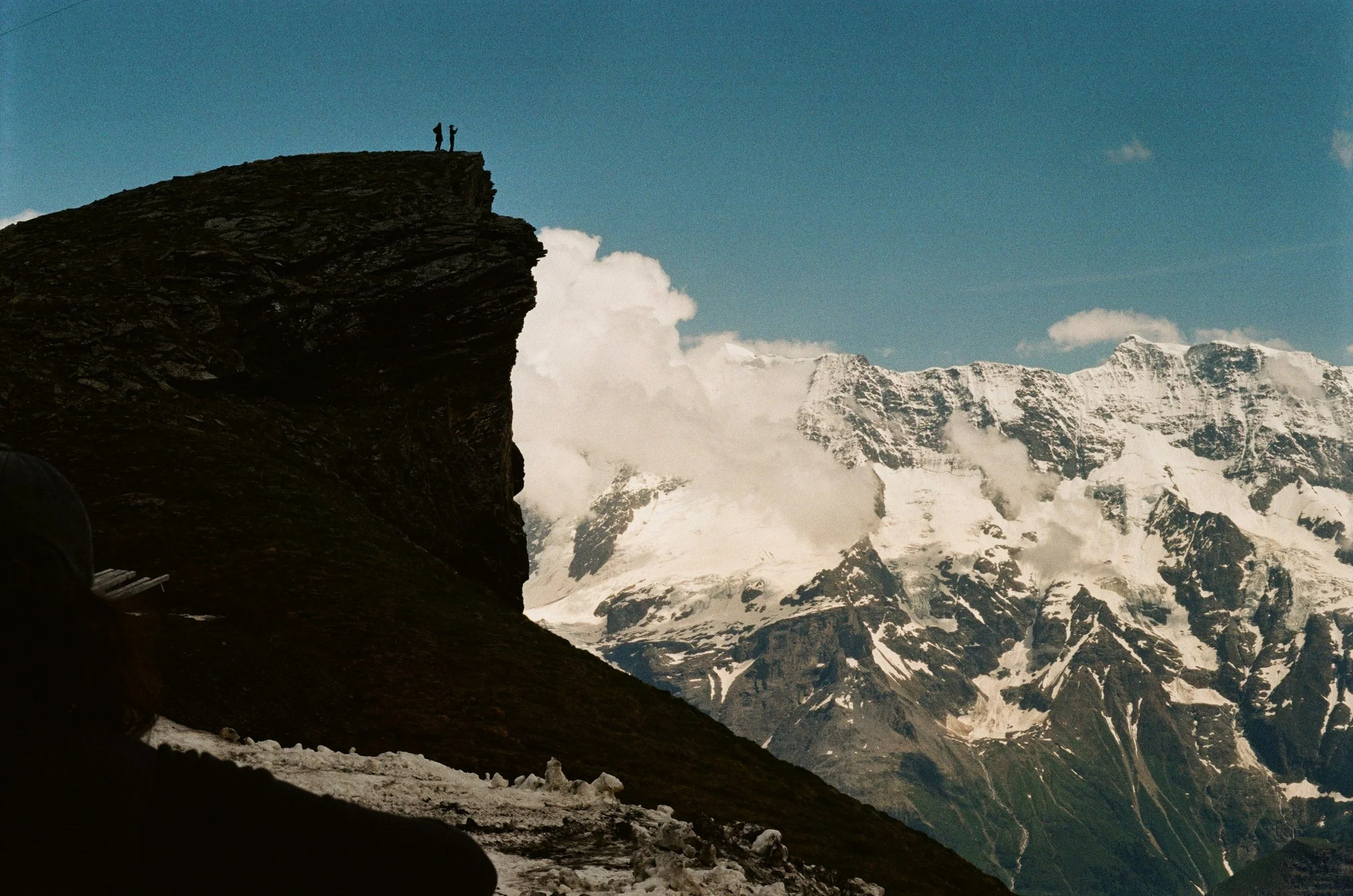 snowy mountains with large cliff in foreground and 2 silhouettes of people standing on its edge