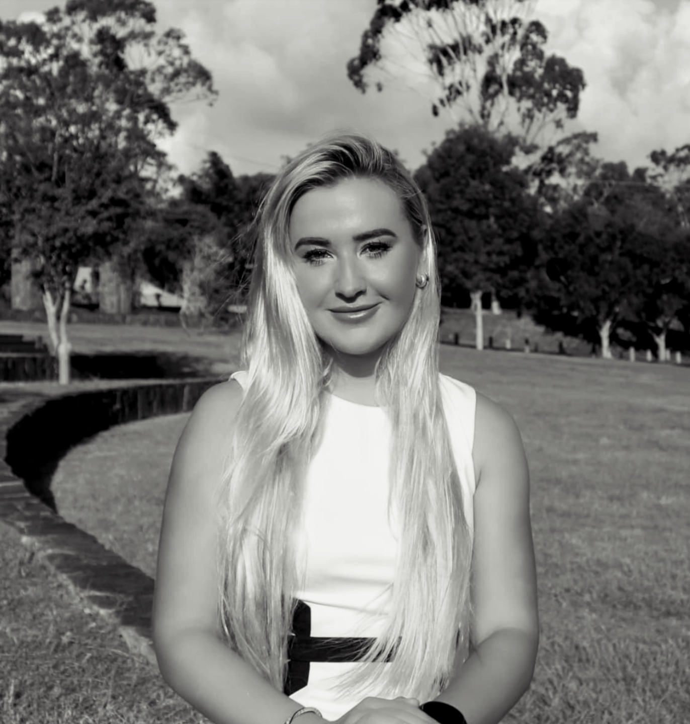 Black and white photo of a young woman with long, wavy hair standing outdoors in a park, with trees and a curved brick wall in the background. Photo of Gabrielle-Beth Volovsky, psychotherapist, counsellor and performance coach.