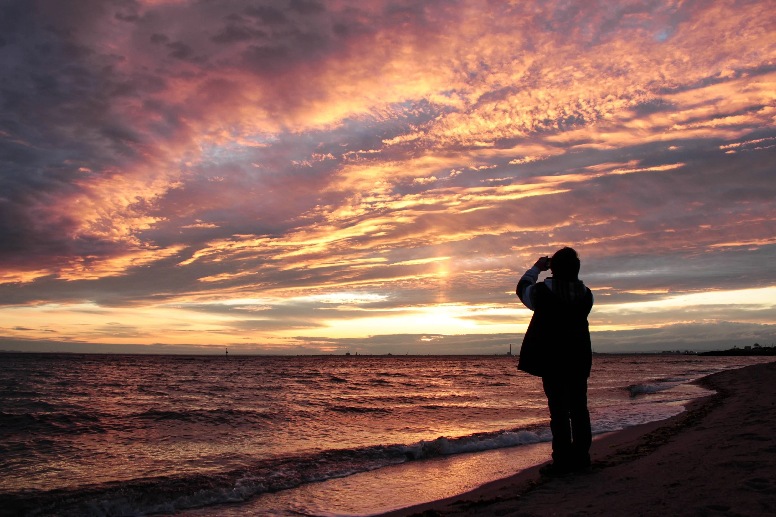 photography of boy standing watching a beautiful pink yellow orange and purple sunset at the beach with water crashing on the shore