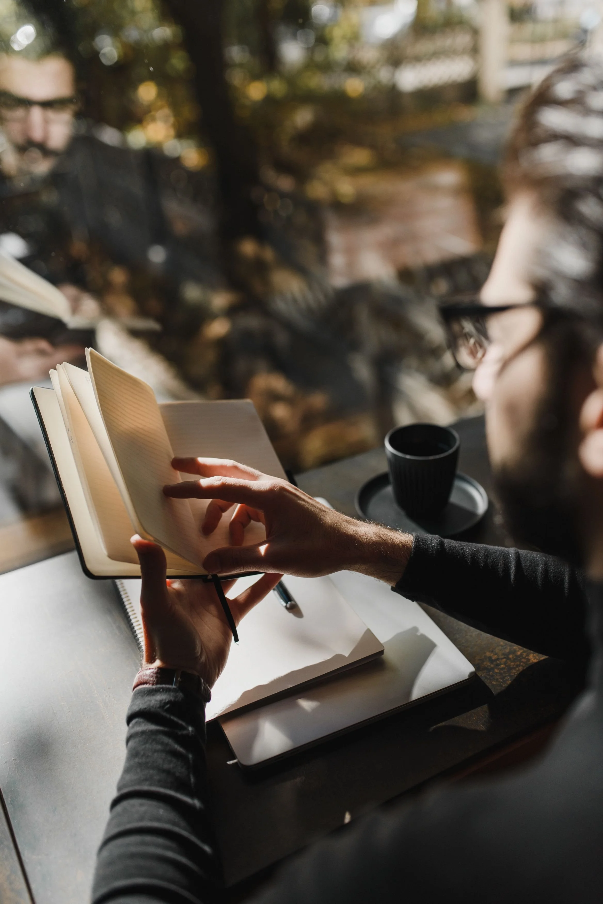 man wearing black reading and writing in notebook and journal, wearing glasses next to reflective window