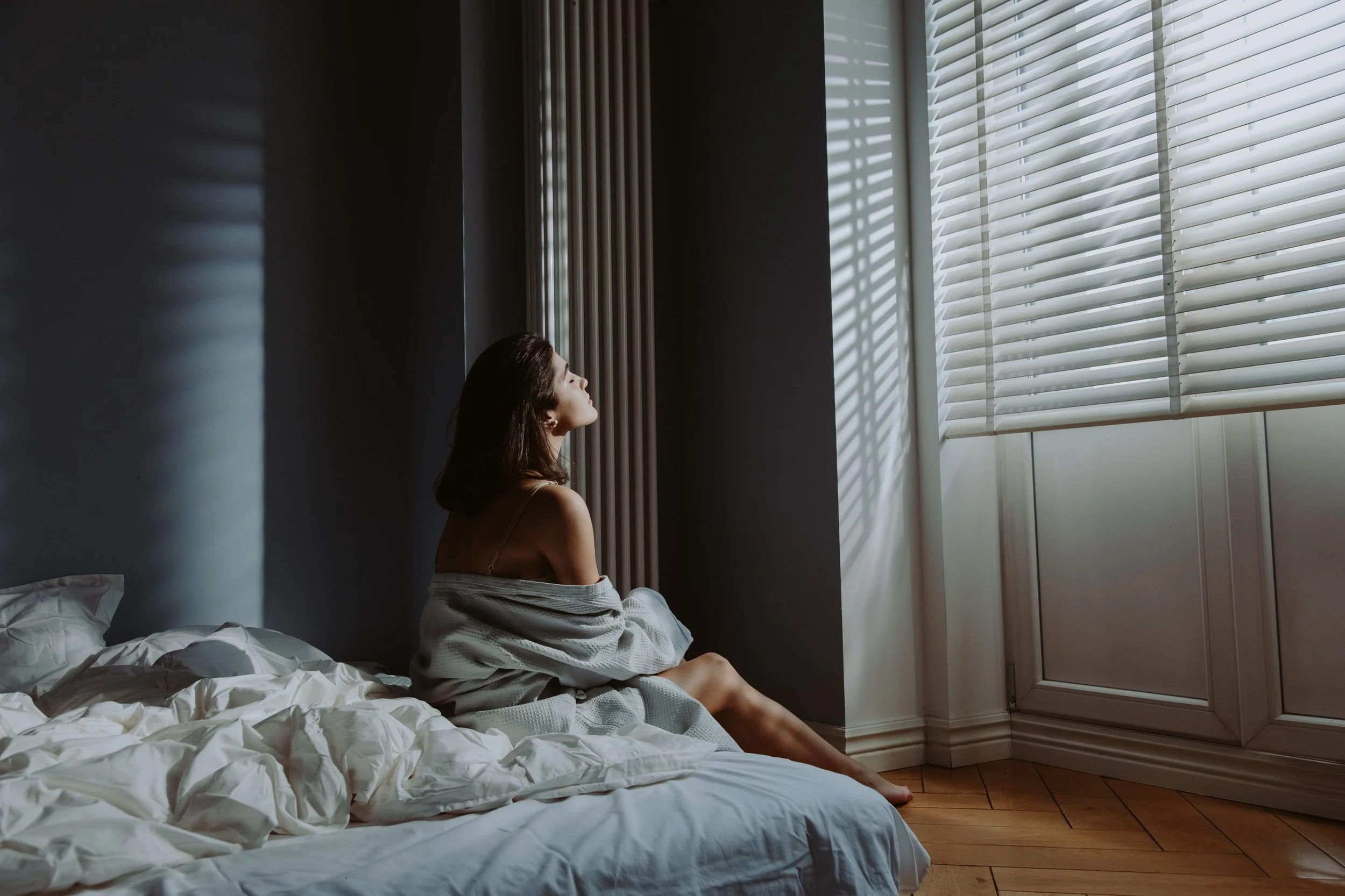 woman waking up in the morning putting her face in the sunlight coming through the blionds on the window whilst sitting on her bed looking cosy and snug in her bedding and blanket