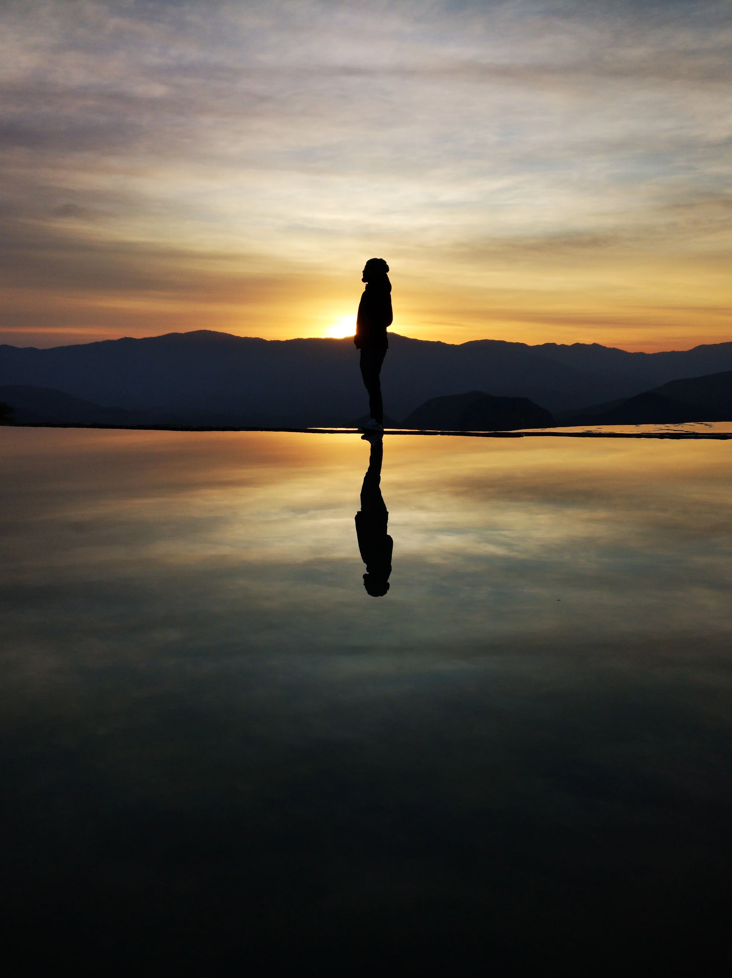 silhouette of person at sunset with beautiful sky colours standing on water with mountains in background and reflection of shadow