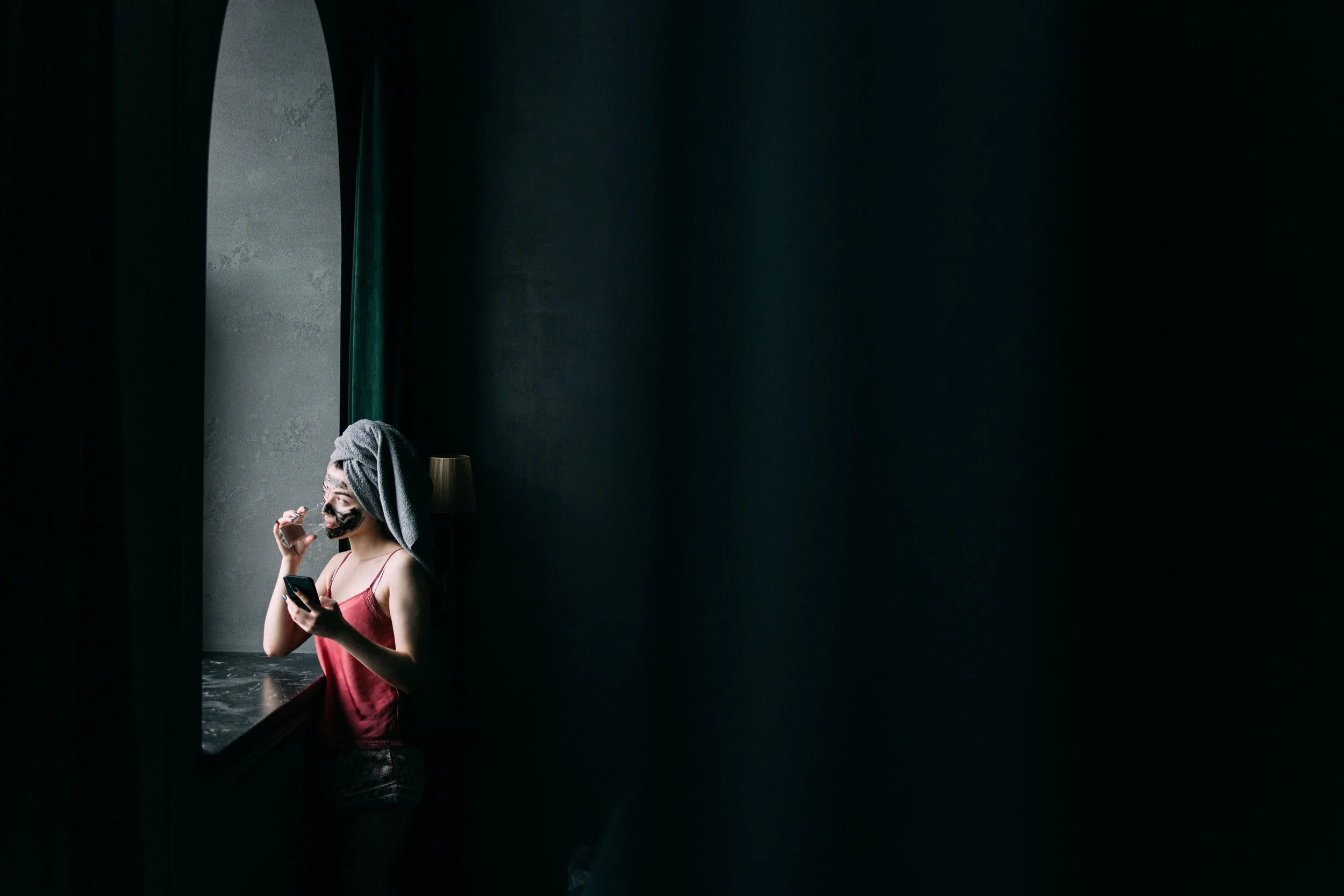 dark room with woman standing in corner by window with clay on her face, towel on head, drinking vitamins and supplements whilst on her phone - wellnessmaxxing