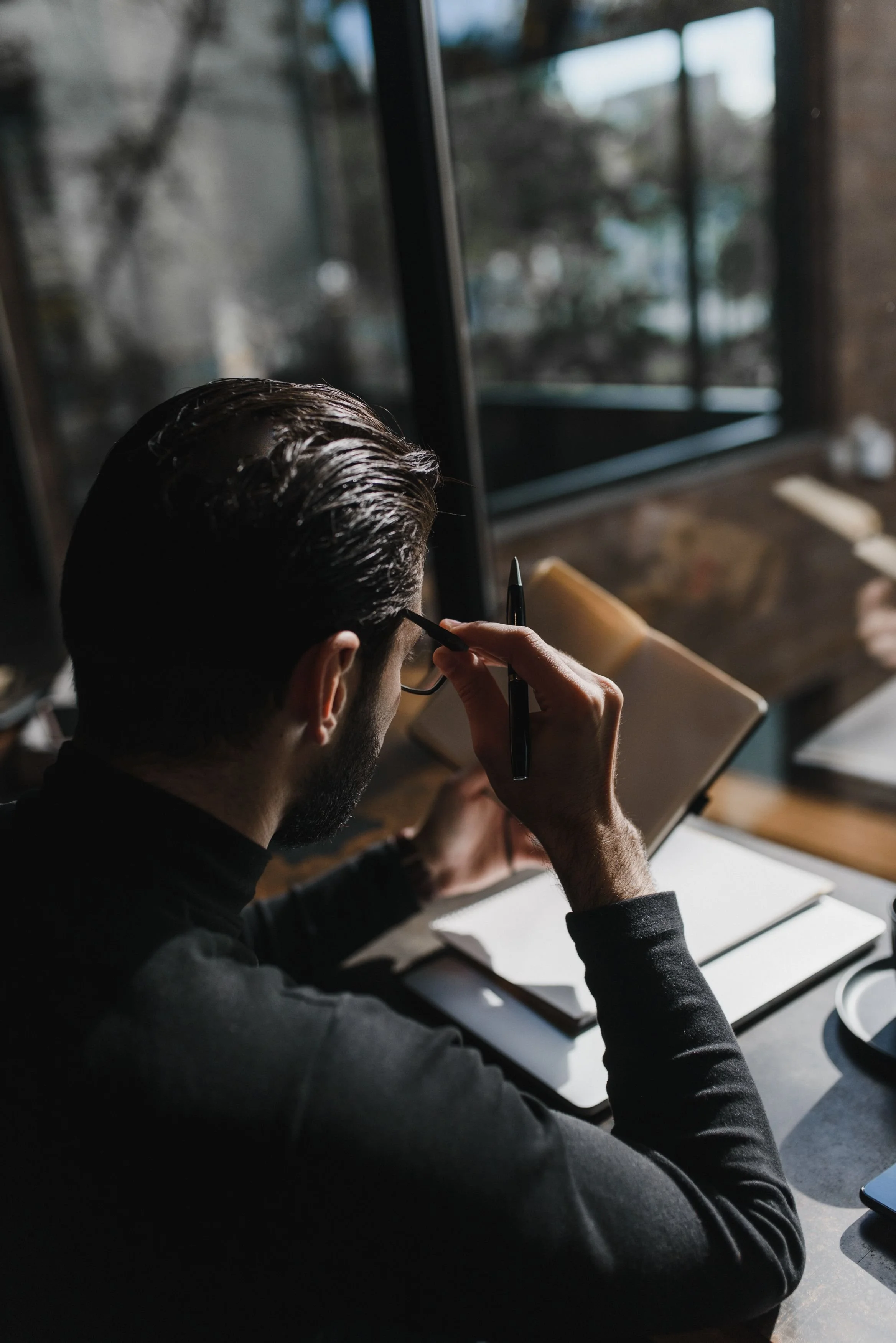man sitting by window, dressed in black, wearing glasses, holding a pen, writing, journalling and reading.