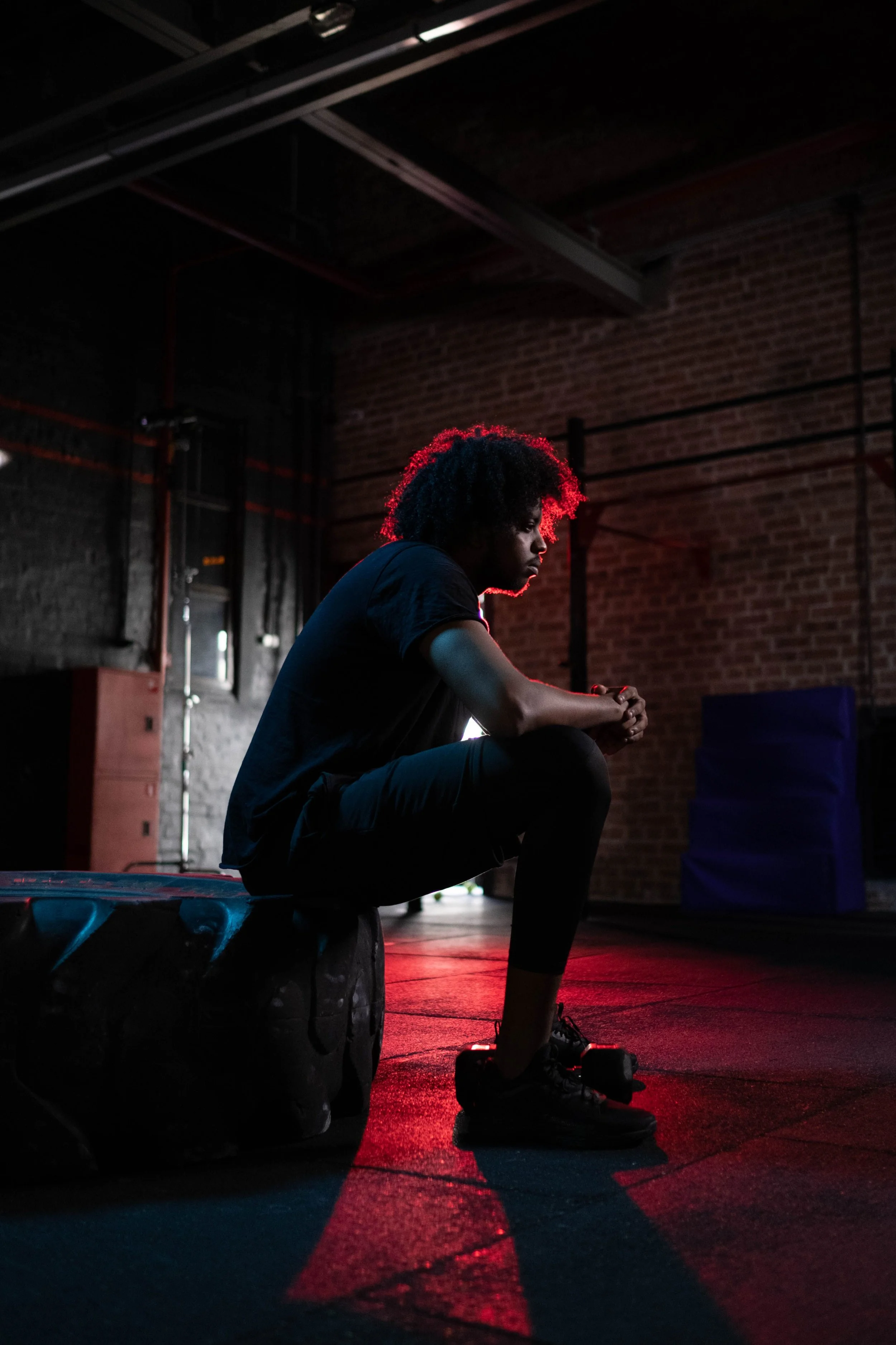 man sitting on tyre at the gym with red light behind him looking pensive and sad unable to explain his feelings, looking burntout