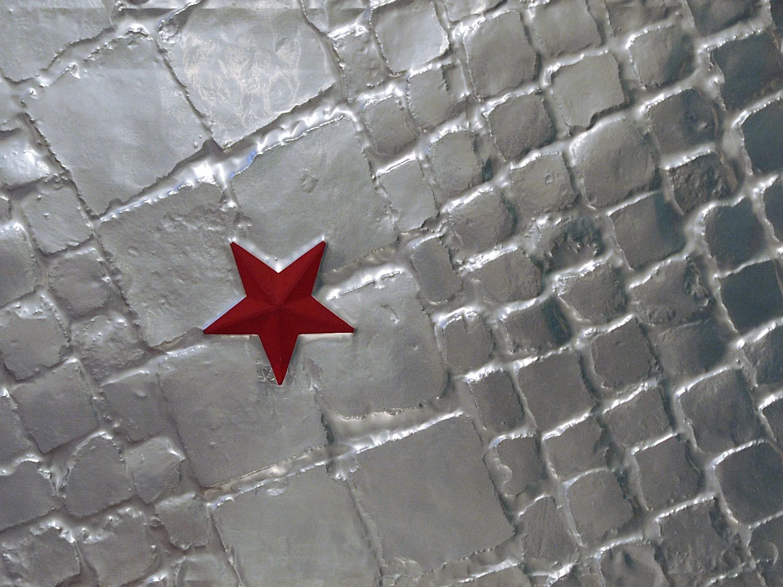 Close-up of a textured gray stone or tile surface with a red star-shaped decoration attached. Baldo Diodato.