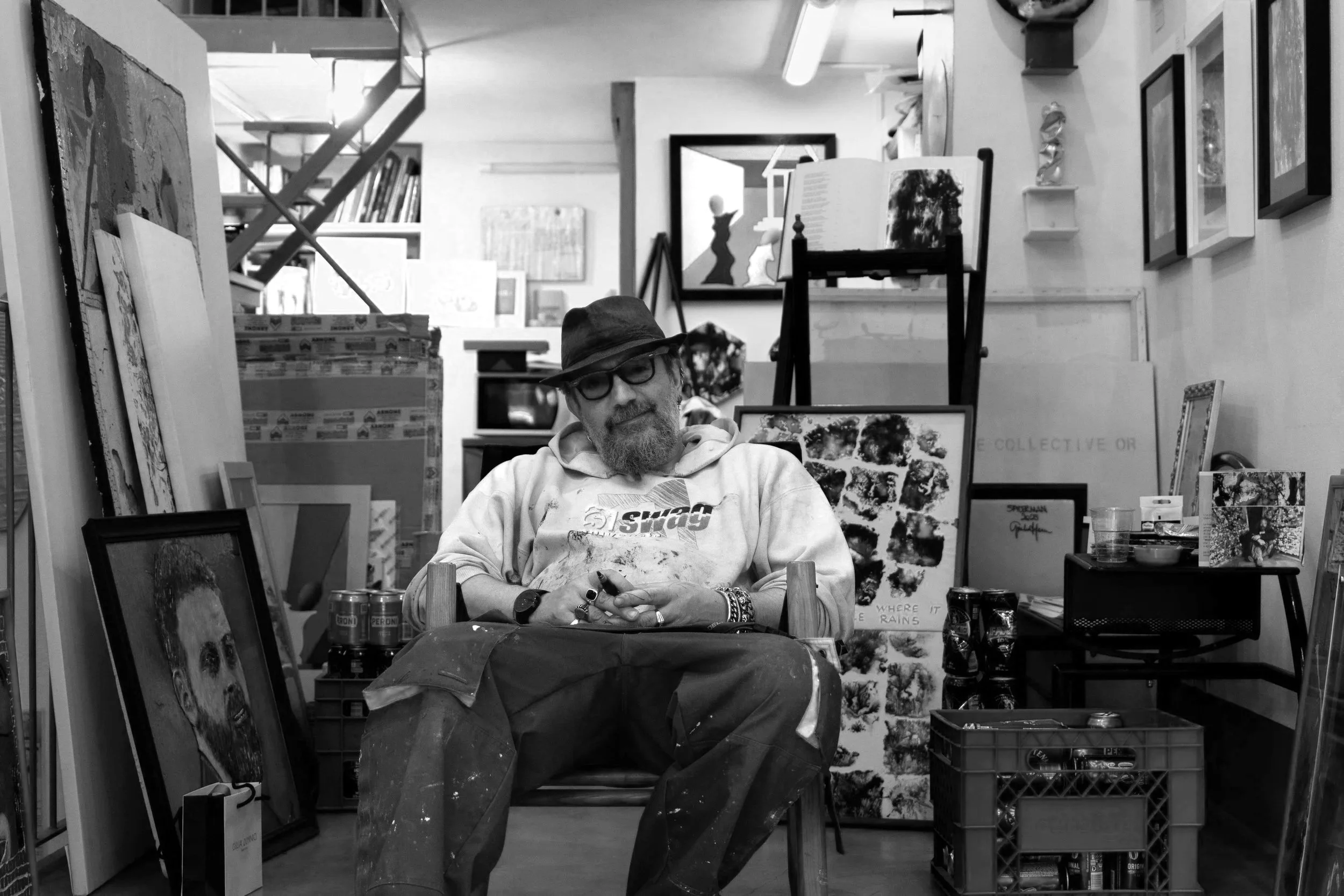 A man with glasses, a beard, and a hat sits in a cluttered art studio, surrounded by paintings, framed artwork, and art supplies.