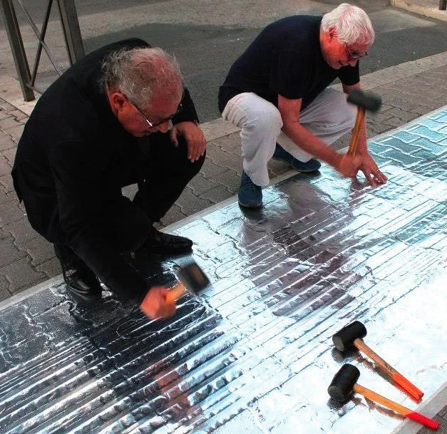Two men are working together to smooth and flatten a large sheet of shiny, metallic material on the ground outdoors, using small hand tools and a roller. Baldo Diodato.