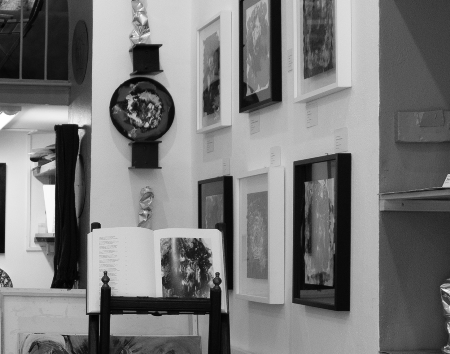 Black and white photo of an art gallery wall displaying various framed abstract paintings. A book open on a stand in front, showing an abstract artwork. The gallery has decorative wall pieces and a small shelf. Some artworks have labels below them.