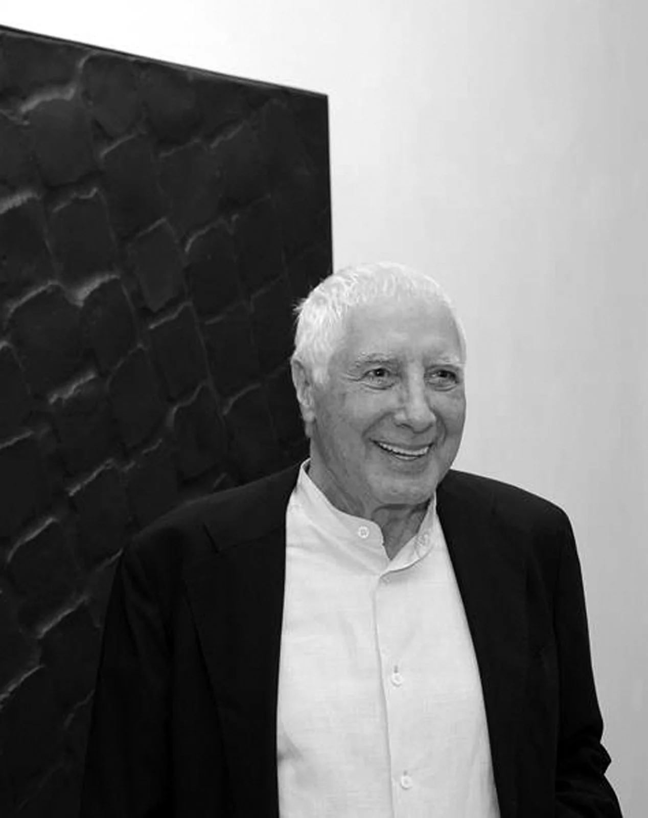 Smiling elderly man with white hair wearing a white button-up shirt and black blazer, standing in front of a textured wall. Baldo Diodato.