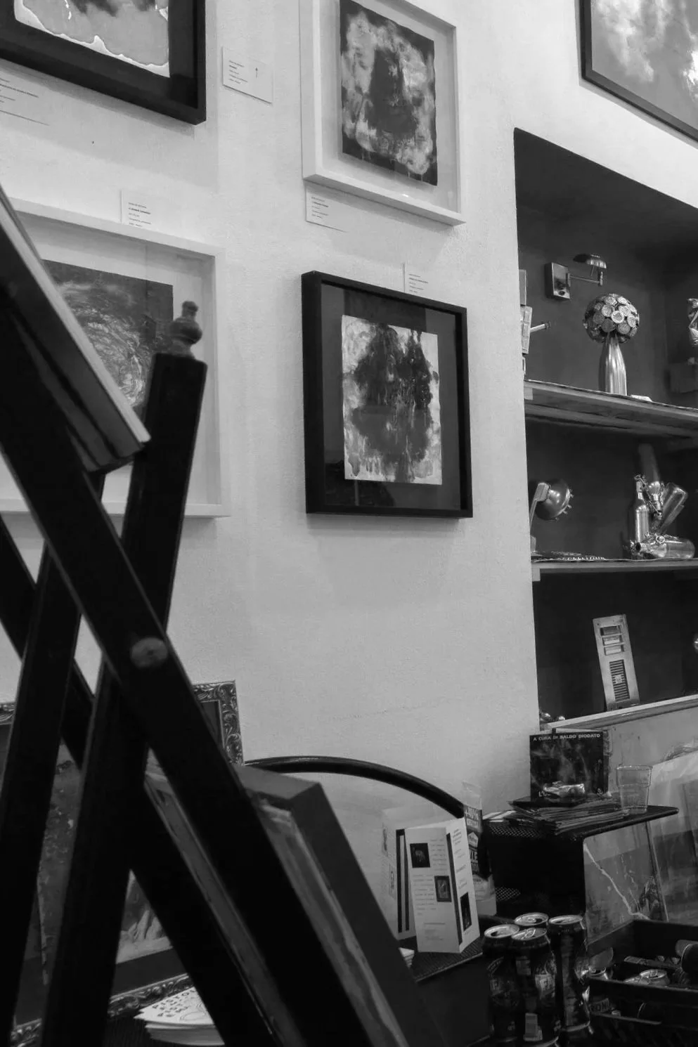 Black and white photo of an interior art gallery with framed paintings on a textured wall, shelves with decorative items, and a table with bottles and boxes.
