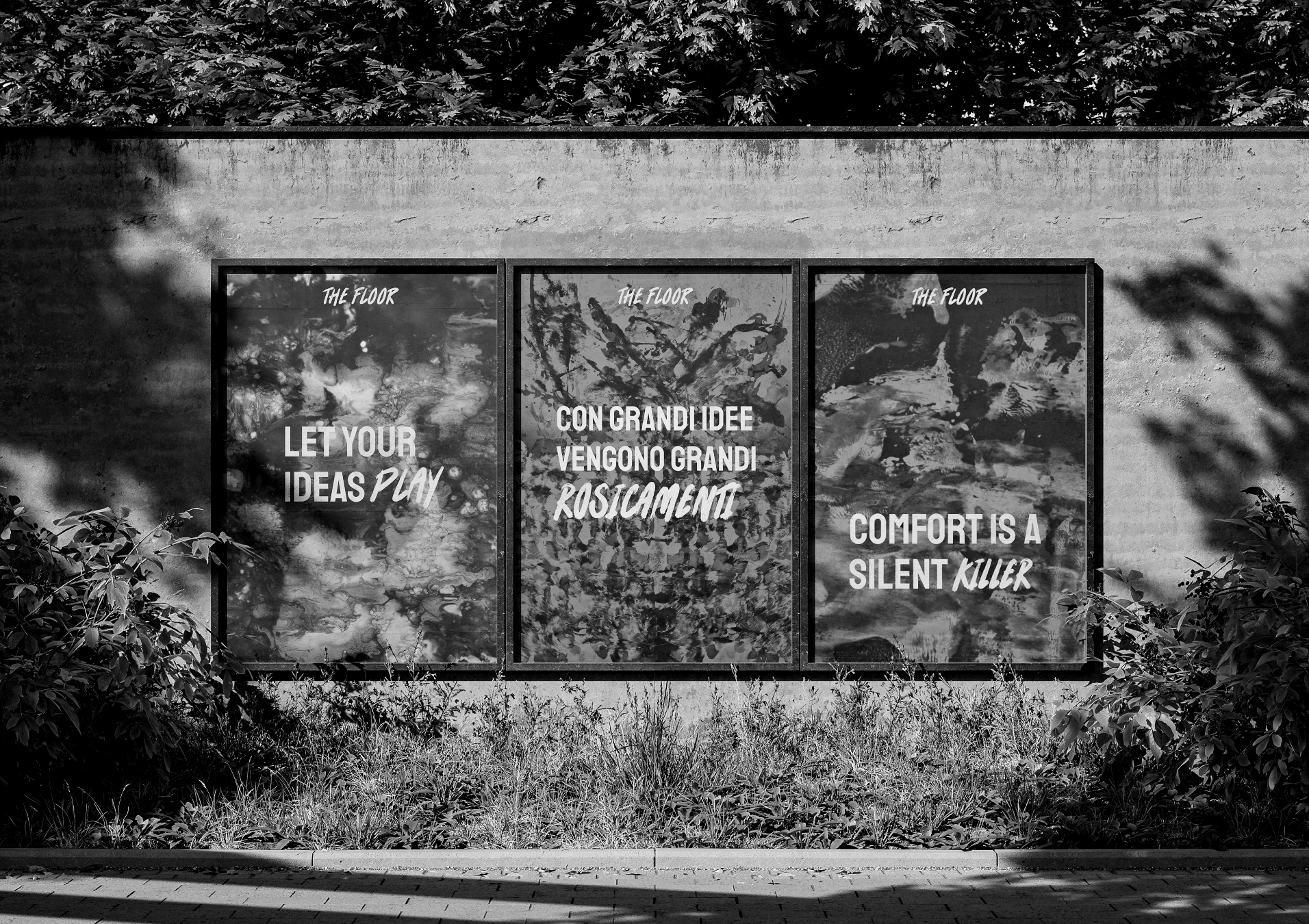 Three large posters on a concrete wall, each with abstract artwork and inspirational quotes about ideas and creativity, surrounded by bushes.