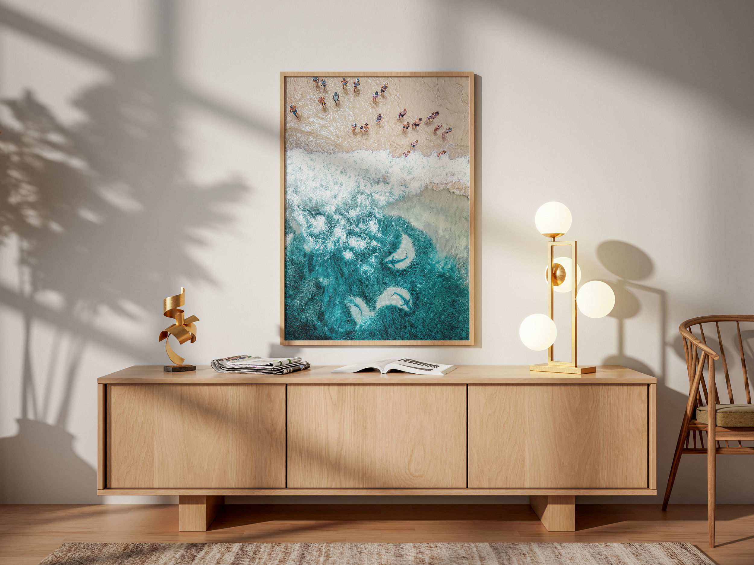 A modern wooden sideboard in a room with minimalist decor, featuring a sculpture, an open book, and a multi-globe table lamp. A large framed artwork of a beach scene hangs above, and a wooden chair is visible to the right. Shadows of plant leaves are