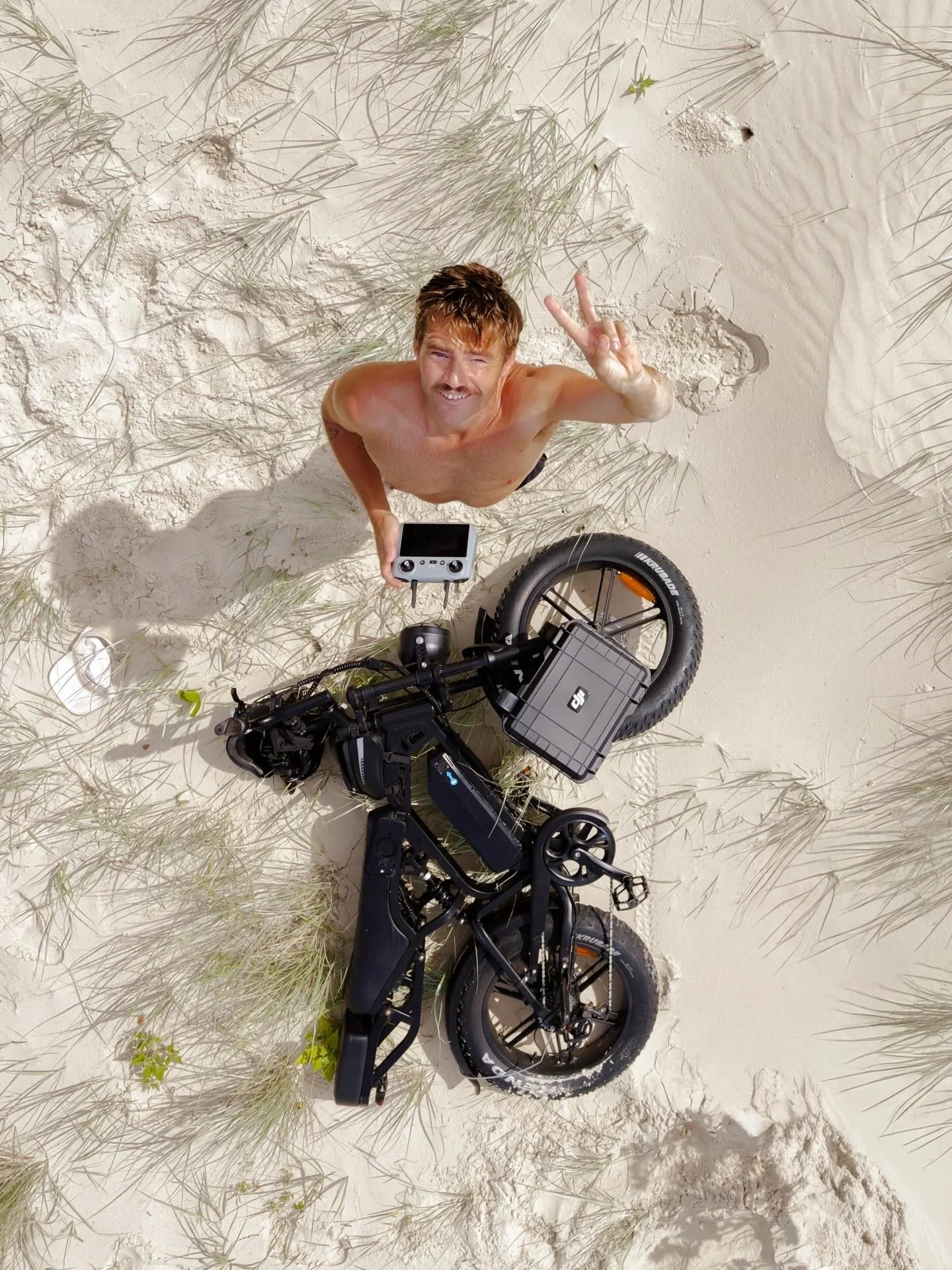 A shirtless man with brown hair making a peace sign while standing next to an electric bicycle on a sandy beach with grass around.