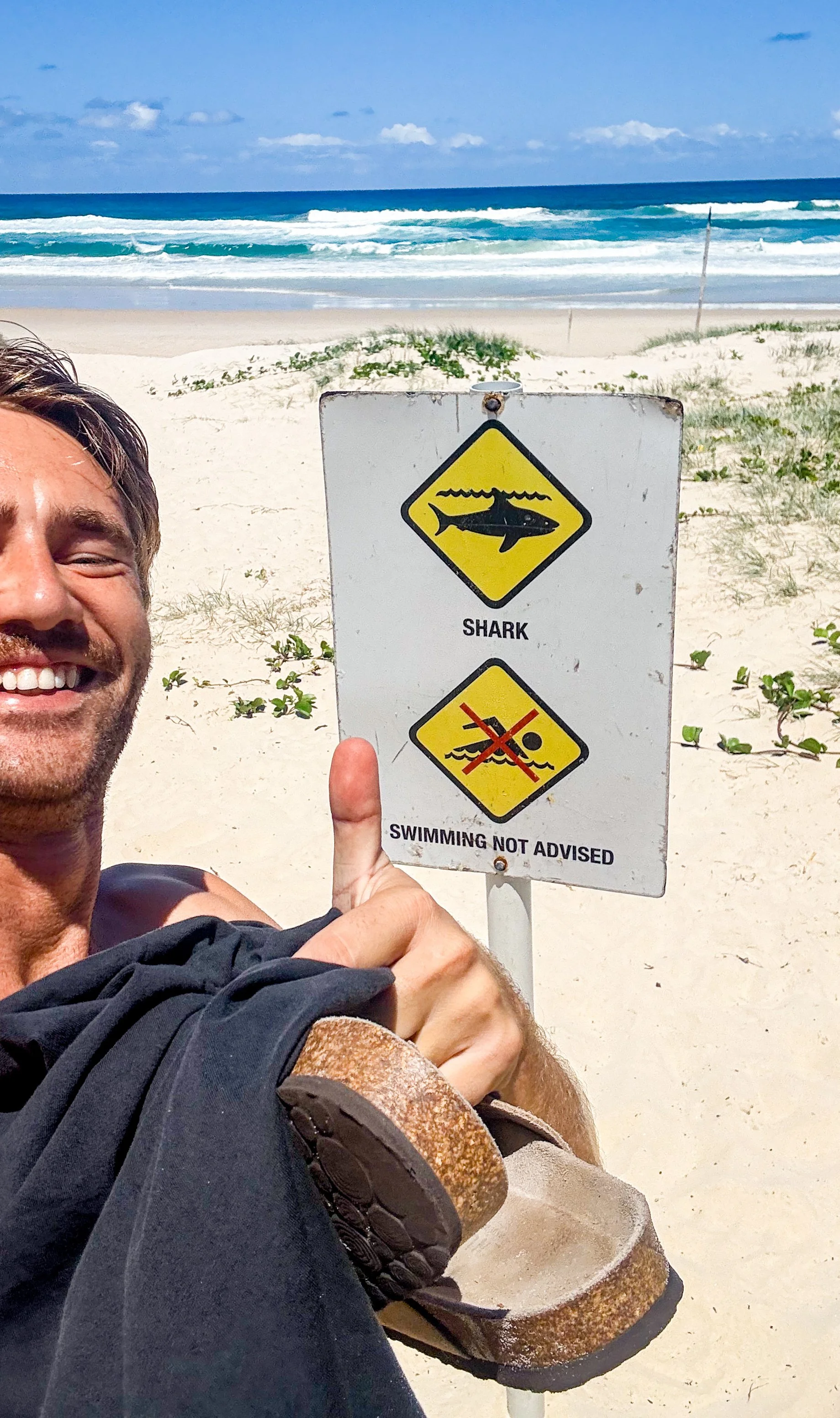 A man smiling on a beach pointing at a sign that warns about sharks and advises against swimming.