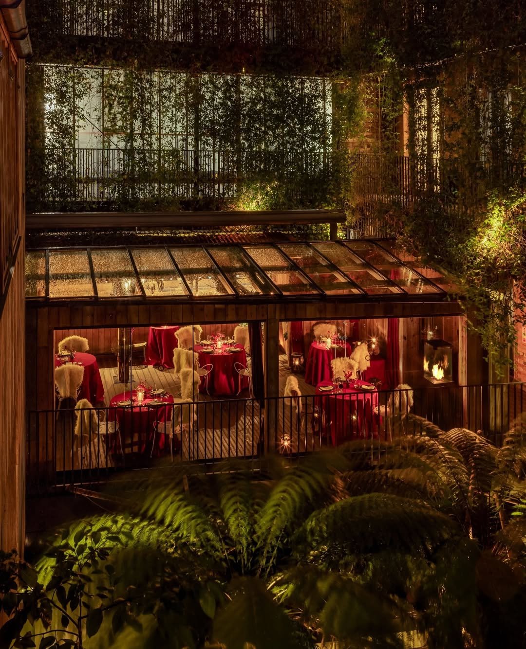 A cozy night view of a glass-enclosed dining area with tables dressed in red tablecloths, surrounded by lush greenery and illuminated by warm lighting.